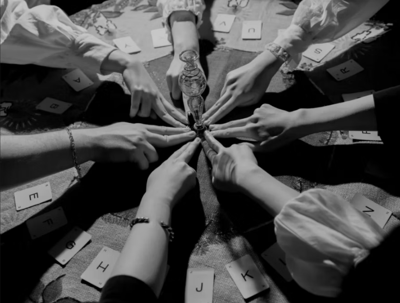 Multiple people place their fingers on a lightbulb centered on a table surrounded by scattered alphabet letter tiles.
