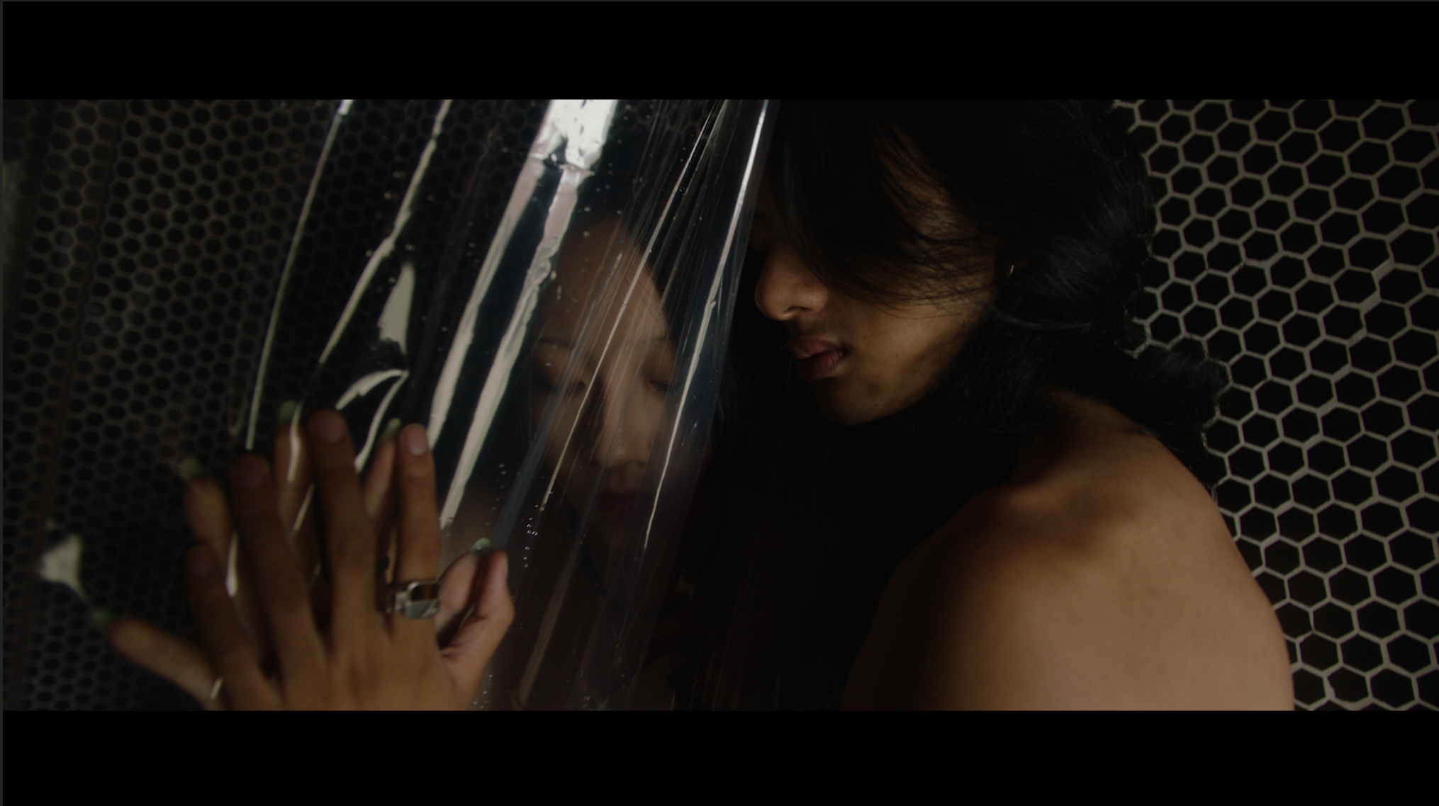 Two women press their foreheads and hands together, separated by a transparent plastic sheet, against a black honeycomb-patterned background, conveying a sense of connection and intimacy.