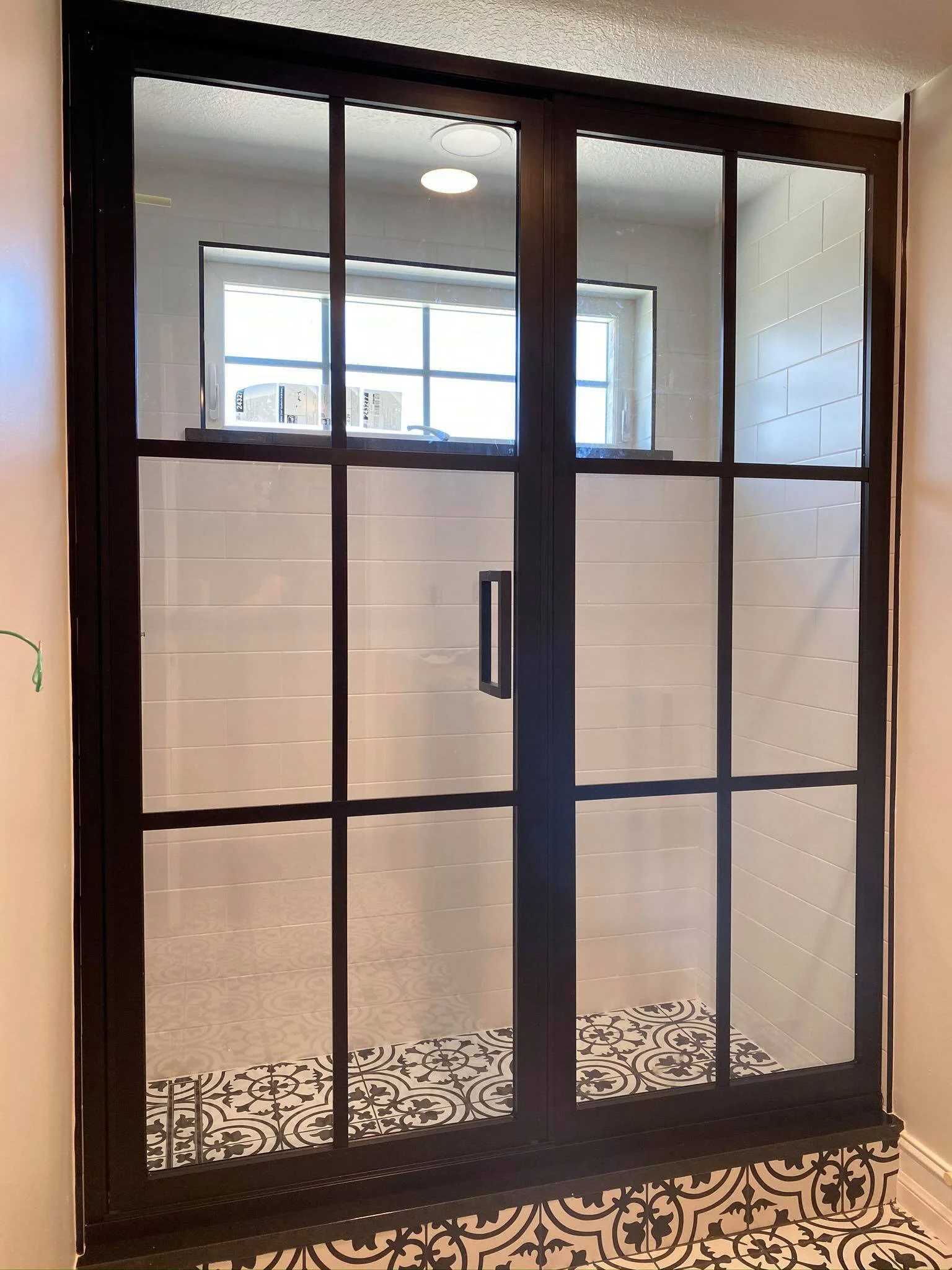 A black metal framed glass shower enclosure with a sliding door, featuring grid-style panes, inside a bathroom with patterned black and white floor tiles.