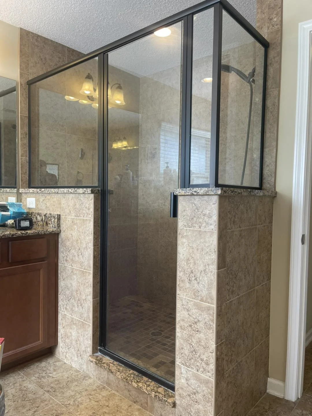 A bathroom shower with glass enclosure, framed with black metal, beige stone tiles on walls and floor, and a granite threshold.