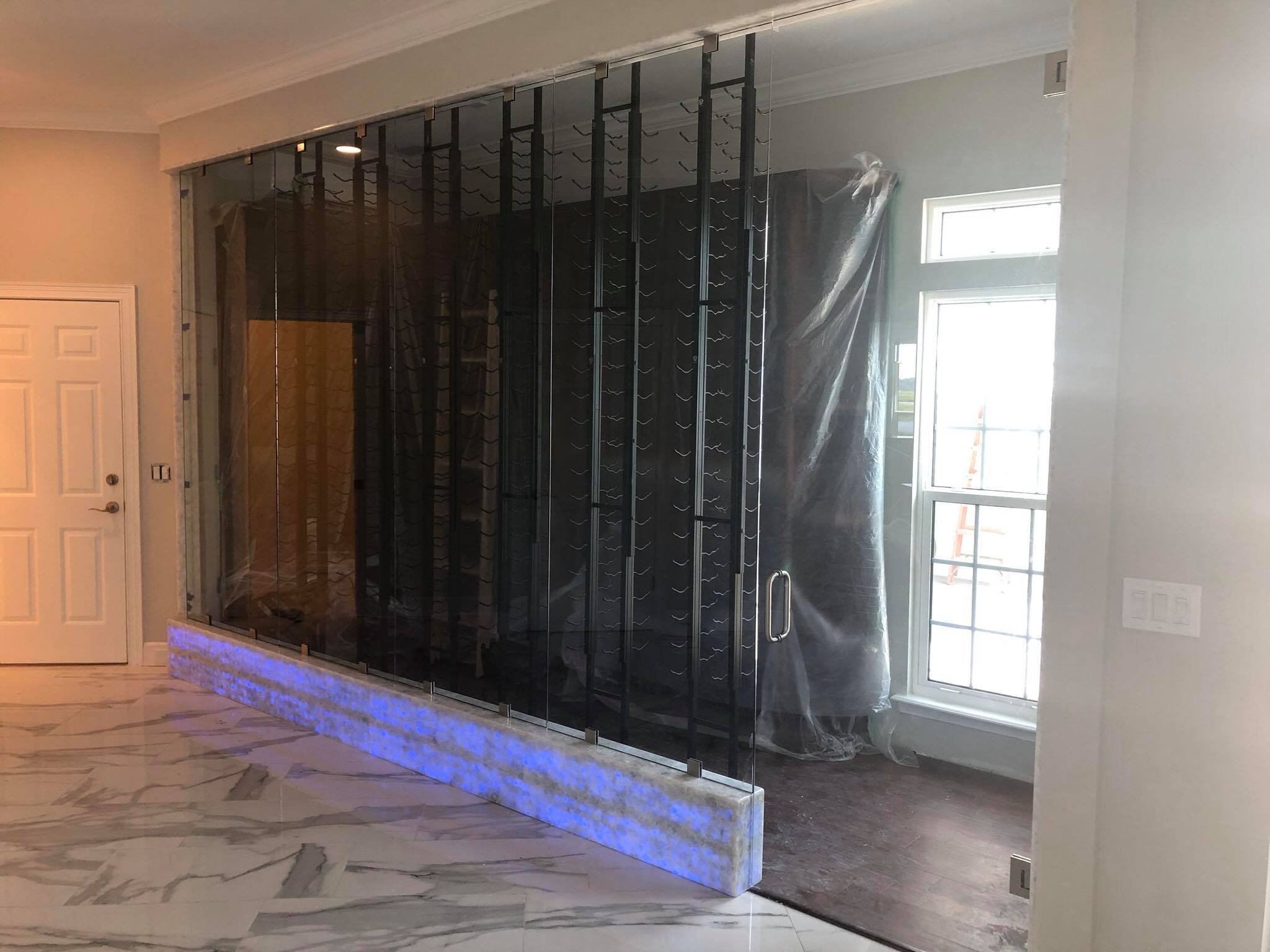 Interior of a house under renovation with a glass-enclosed wine cellar, a marble floor, a white door, and large windows letting in natural light.