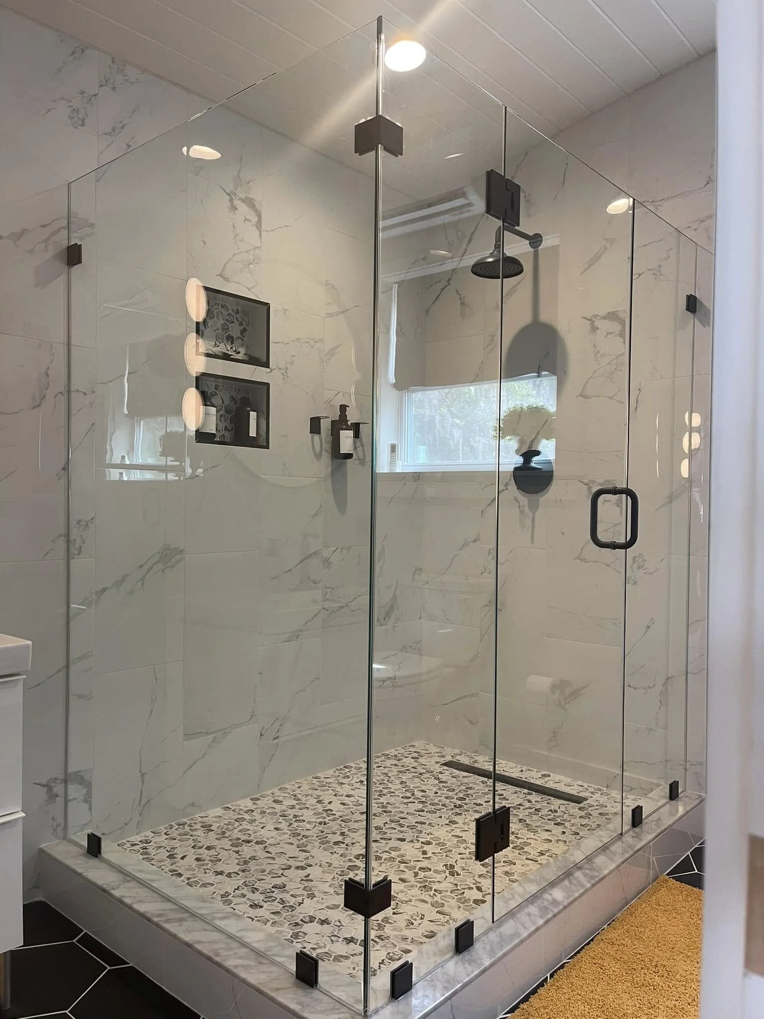 Modern glass-enclosed shower with marble walls, pebble tile floor, black fixtures, and a window with natural light.