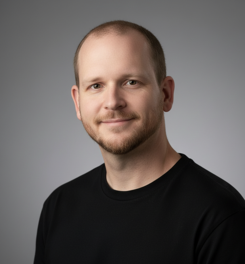 Headshot of a man with short light brown hair and a beard, wearing a black shirt, smiling slightly, against a plain gray background.