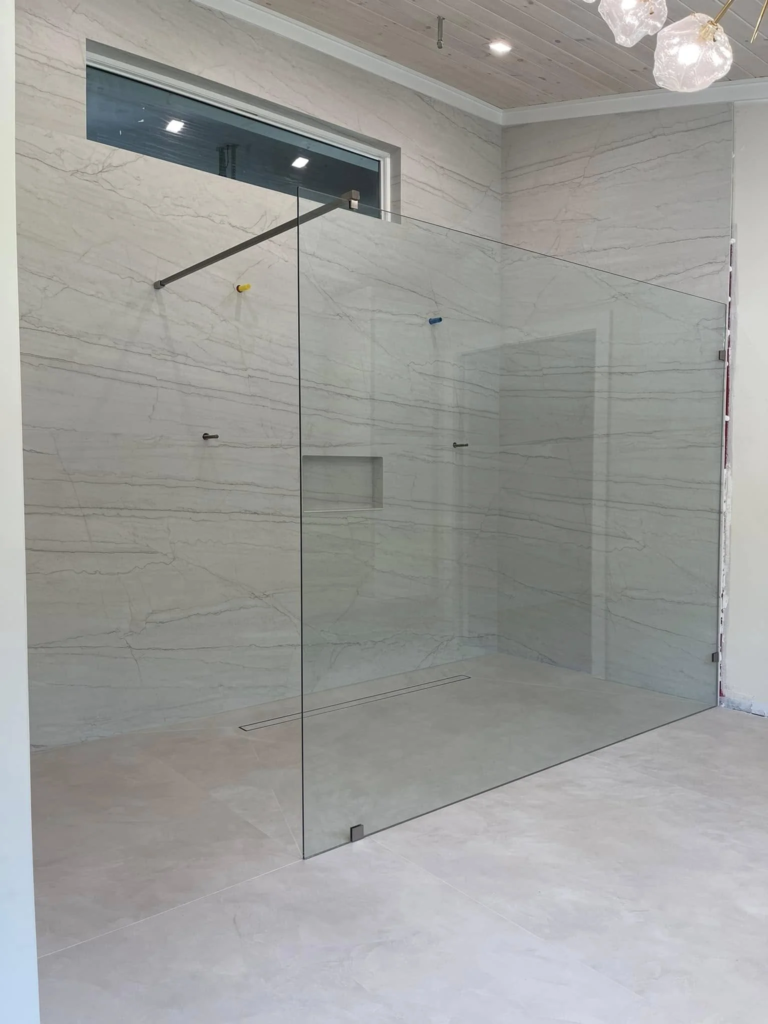 Modern walk-in shower with glass walls, built-in niche, and a window above with a light-colored marble finish.
