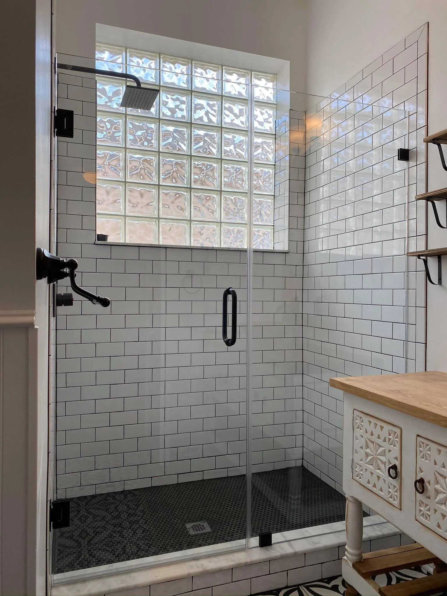 A modern glass-enclosed shower with white subway tile walls, a large window made of glass blocks, and a black rain showerhead. There are wooden shelves on the wall and a rustic vanity nearby.