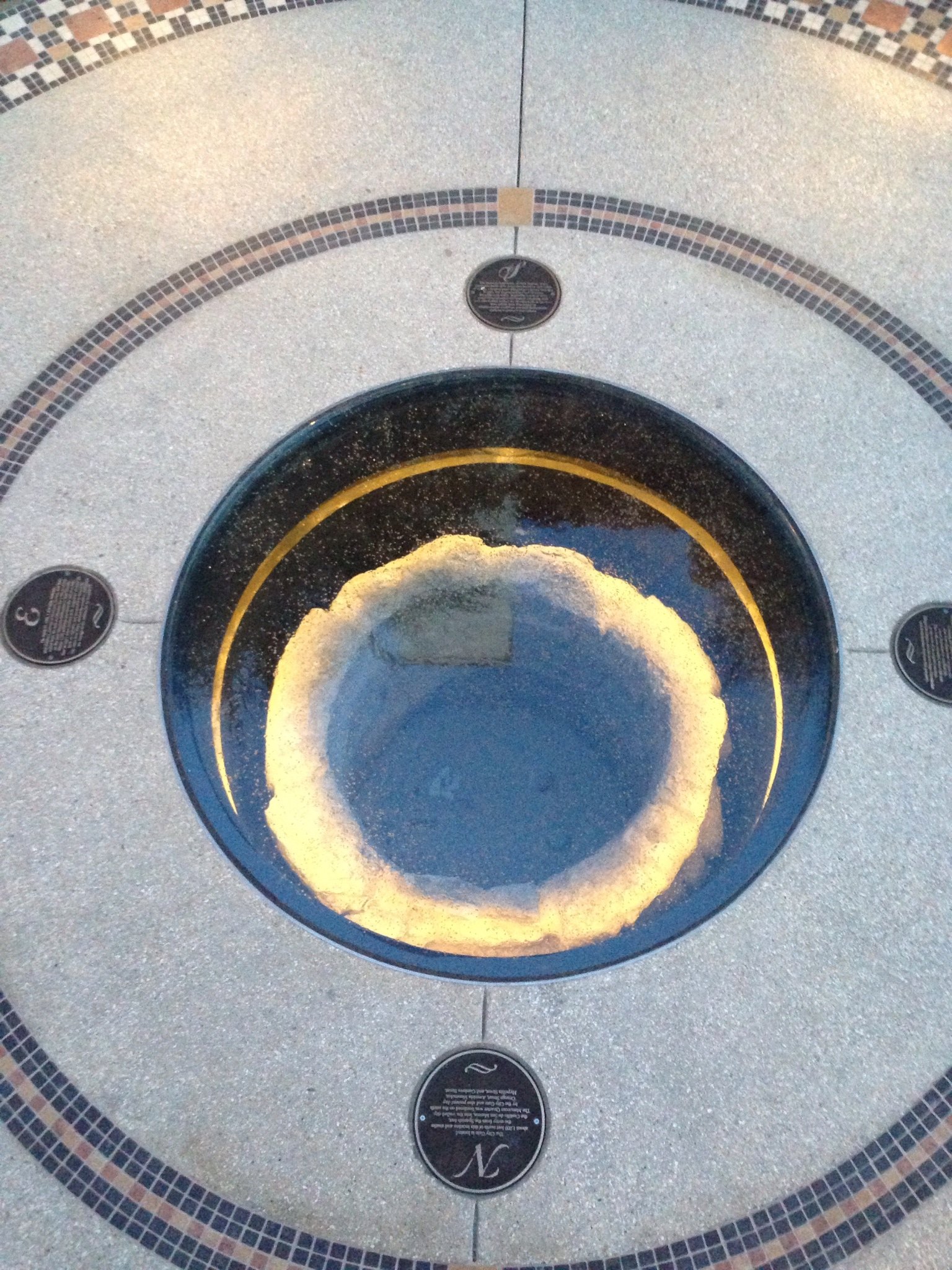 A circular opening in the floor of a building, with surround tiles and four black informational plaques around it, displaying a ring of white sand or powder inside.