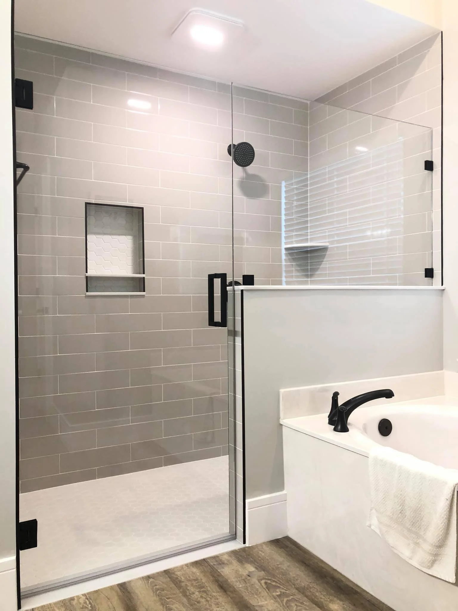 Modern bathroom featuring a glass-enclosed shower with white tiles, a built-in niche, and a black showerhead. Next to the shower is a white bathtub with a black faucet and a towel hanging over the side. The bathroom floor is wood-look vinyl.