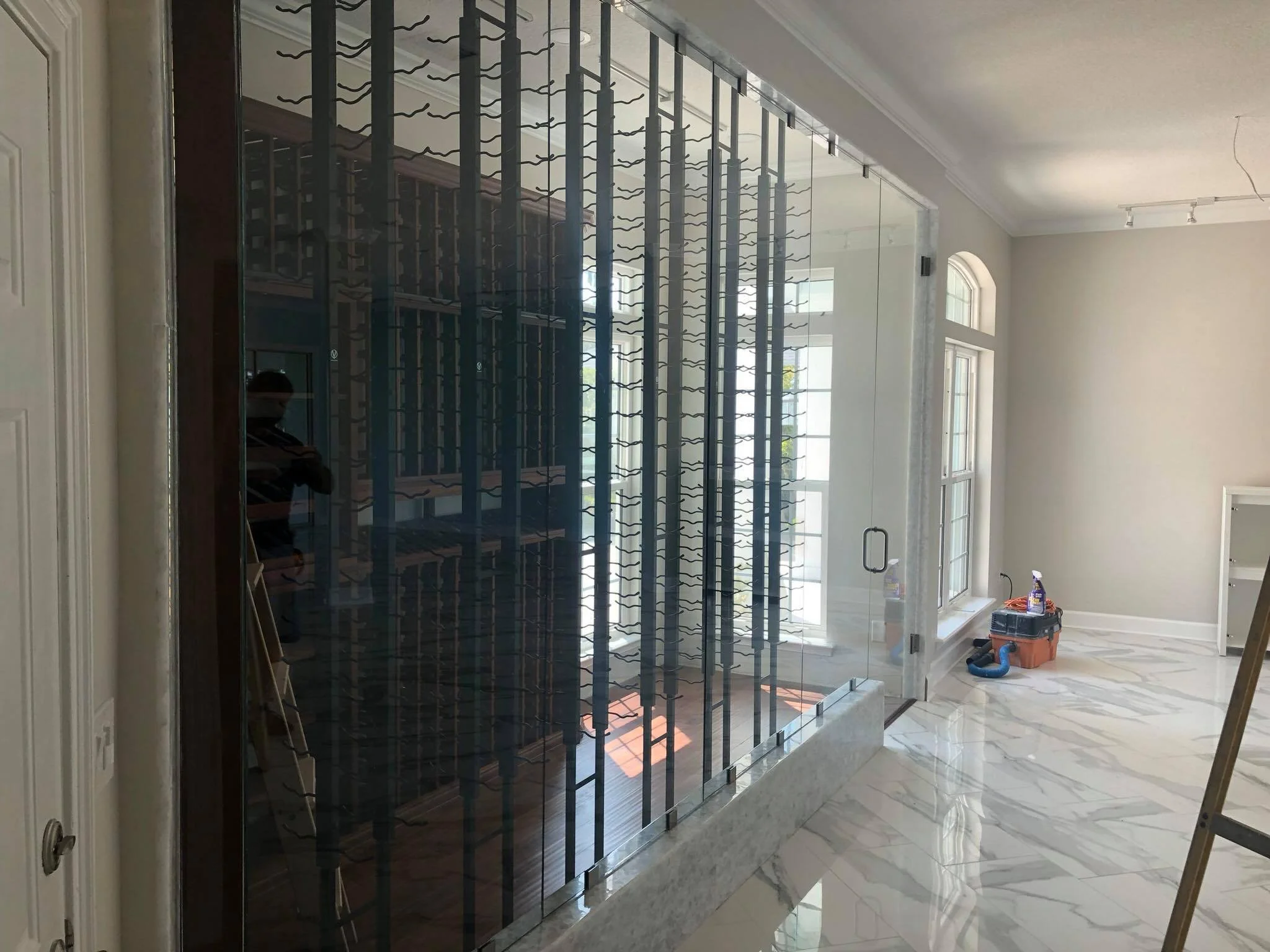 An indoor space with large windows and marble flooring under construction. There is a glass panel with a metal wine bottle holder design, a small white shelf, and a toolbox with cleaning supplies on the floor.