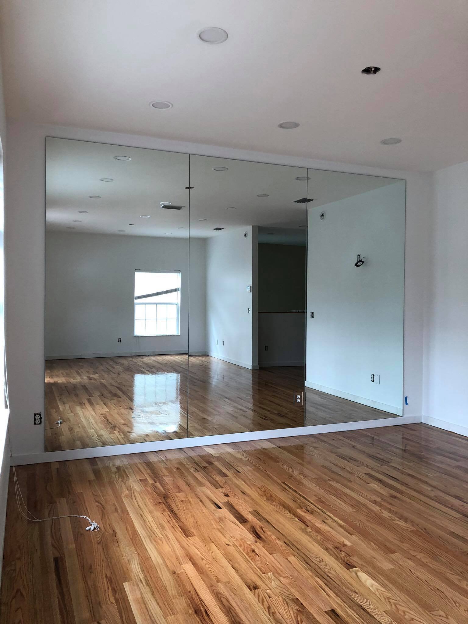 Empty room with hardwood floors, white walls, and a large mirror wall reflecting the space, with electrical outlets and ceiling lights.