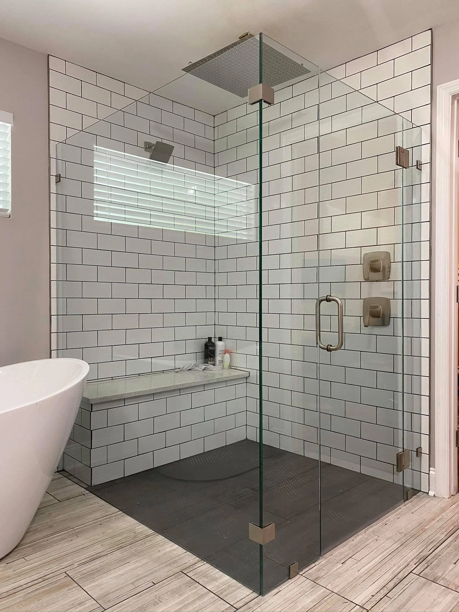 A modern walk-in shower with glass walls, white subway tiles, a wall-mounted rainfall shower head, built-in shelf with toiletries, and wood-look tile flooring outside.