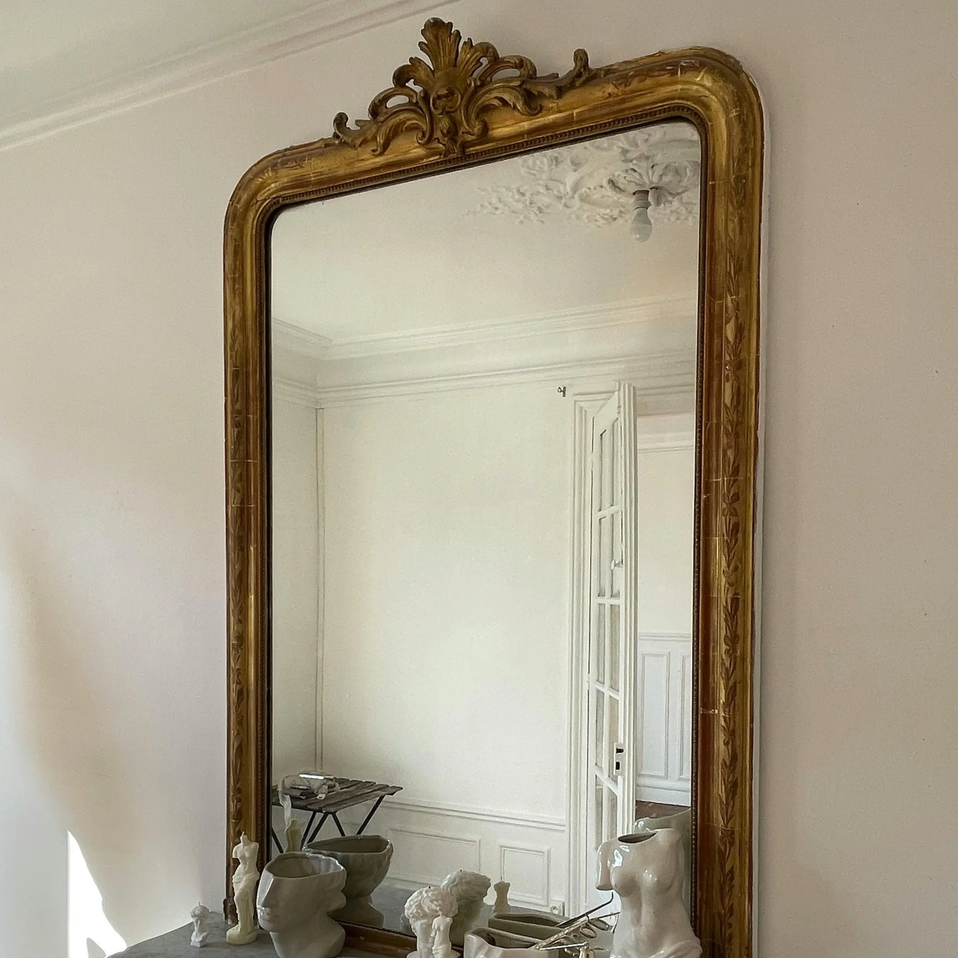 An ornate gold-framed mirror mounted on a white wall, reflecting a room with white walls, decorative molding, and an open door with glass panes.