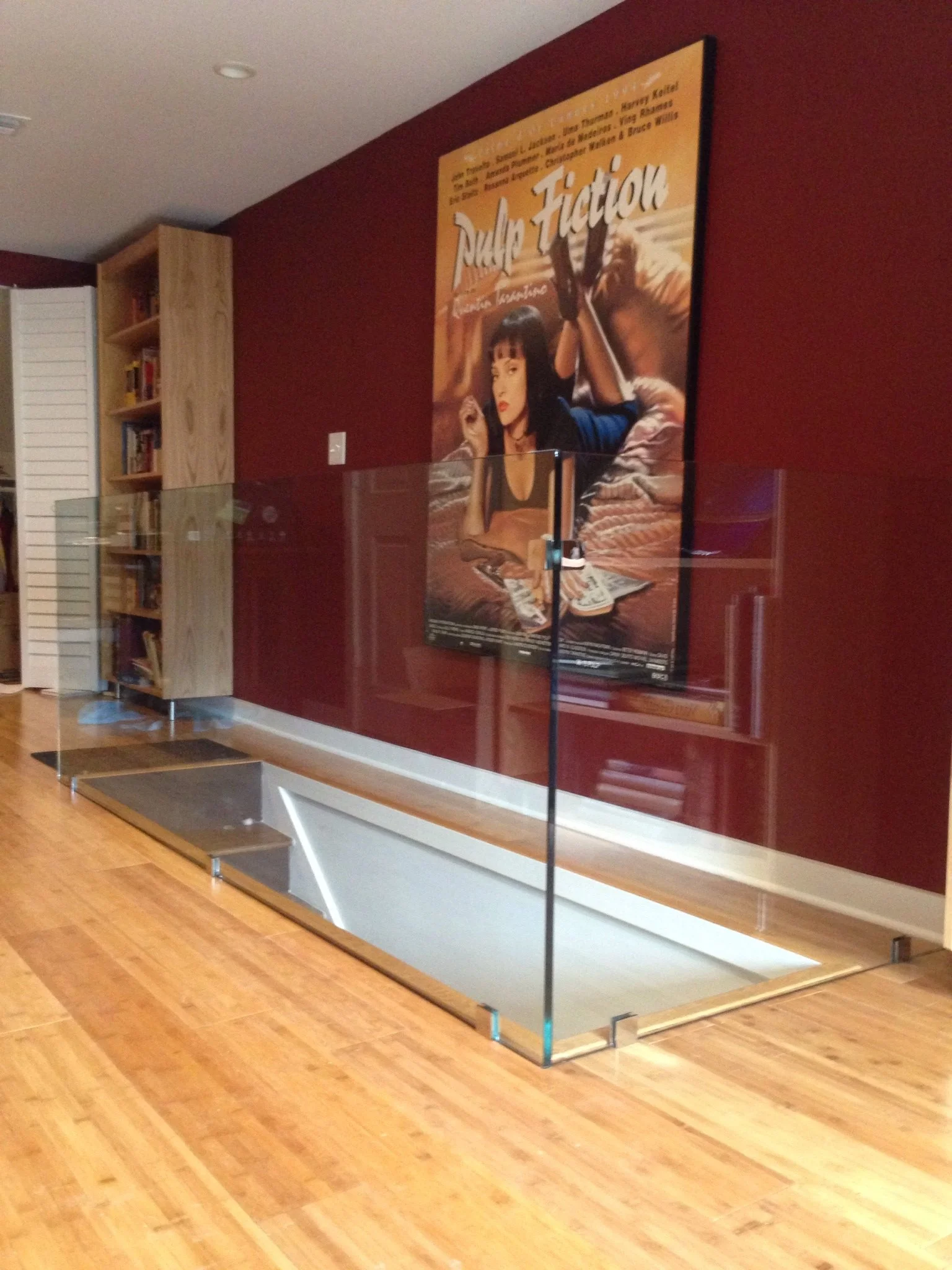 Living room with a large movie poster on a red wall, an open staircase with glass panels, a bookshelf, and wooden flooring.