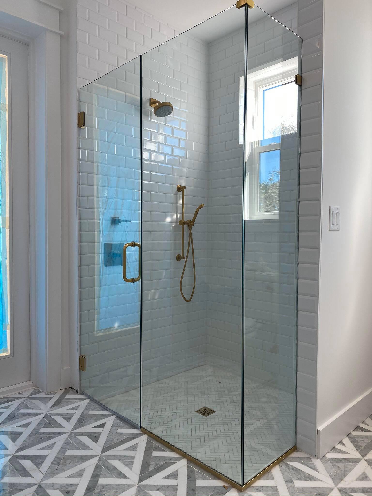 A modern glass shower enclosure with brass fixtures in a bathroom with white subway tiled walls and patterned tile floor.
