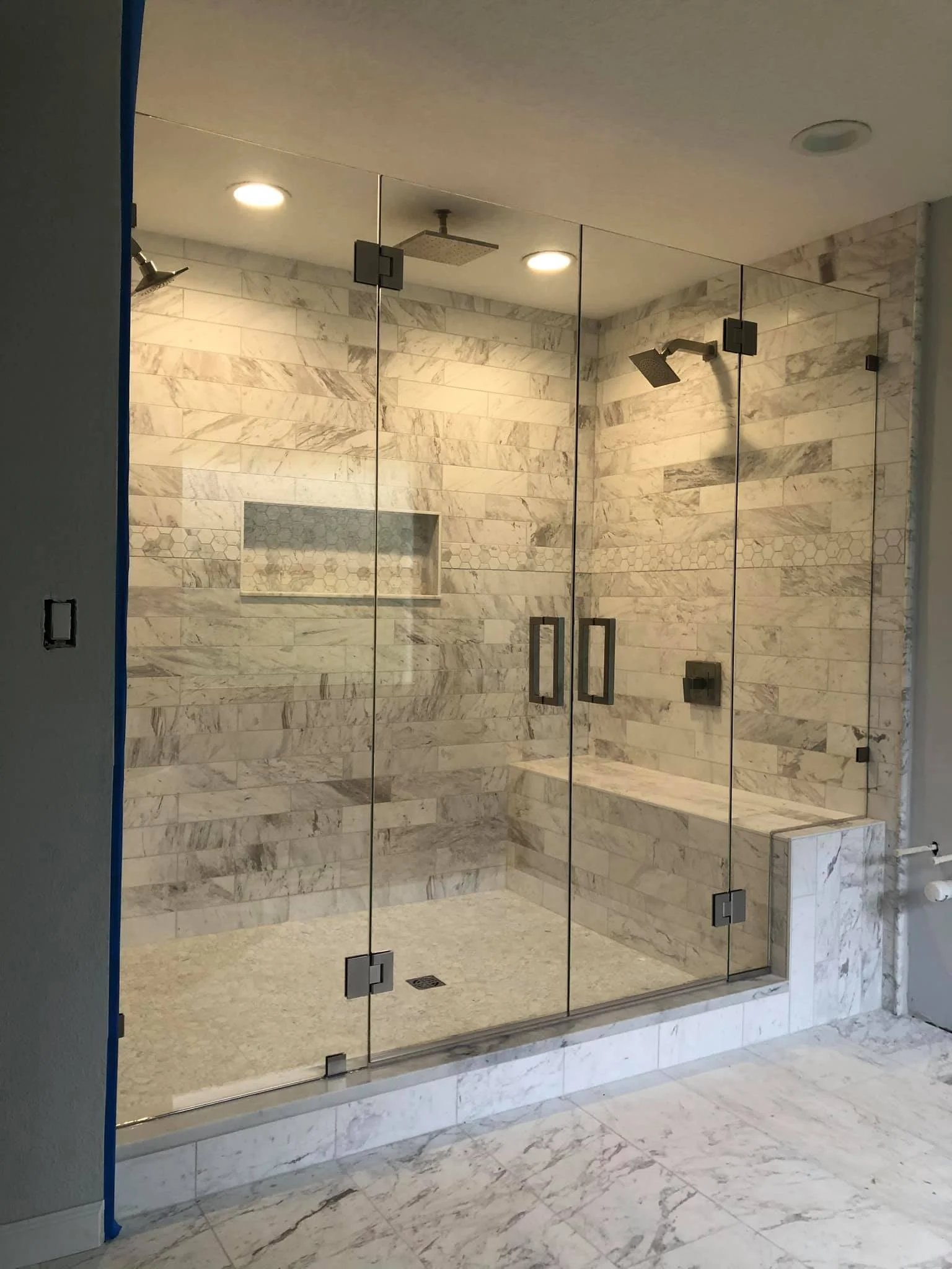A modern walk-in shower with glass doors, marble walls, and a built-in marble bench. It has two overhead shower heads and a small niche in the wall for storage.