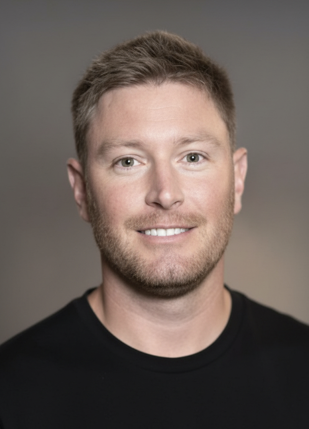 Headshot of a young man with short light brown hair and a beard, smiling, wearing a black shirt, with a neutral gray background.