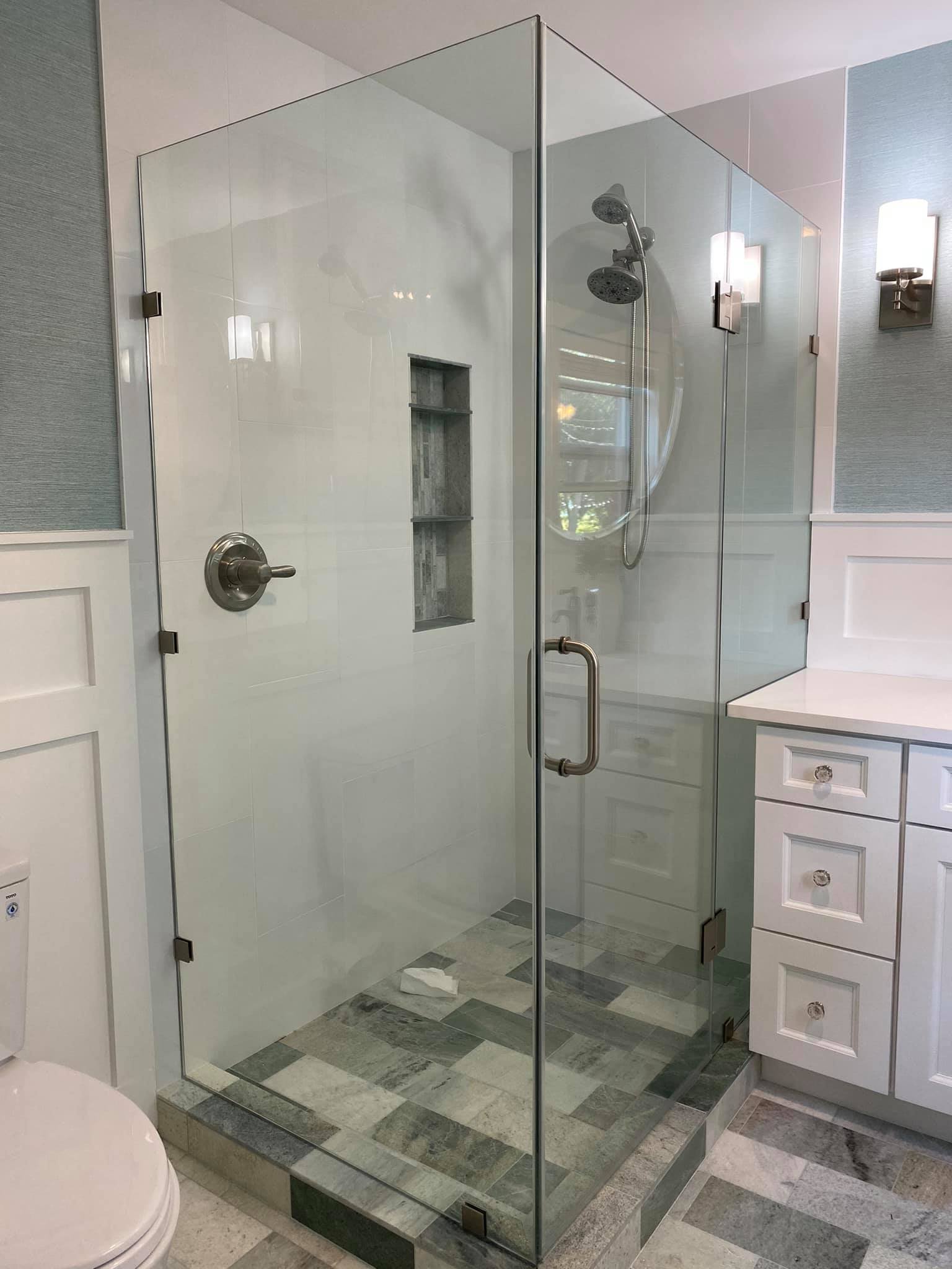 Modern glass shower enclosure with chrome fixtures, built-in shelving, and multiple showerheads in a bathroom with white cabinetry and wall-mounted lighting.