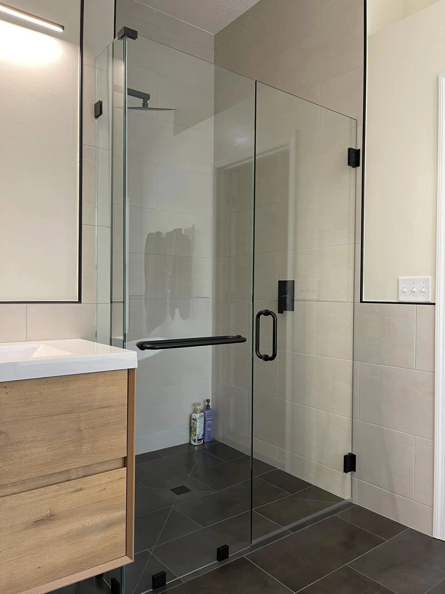 Glass shower enclosure with black hardware in a modern bathroom, with soap and shampoo bottles on the floor inside.