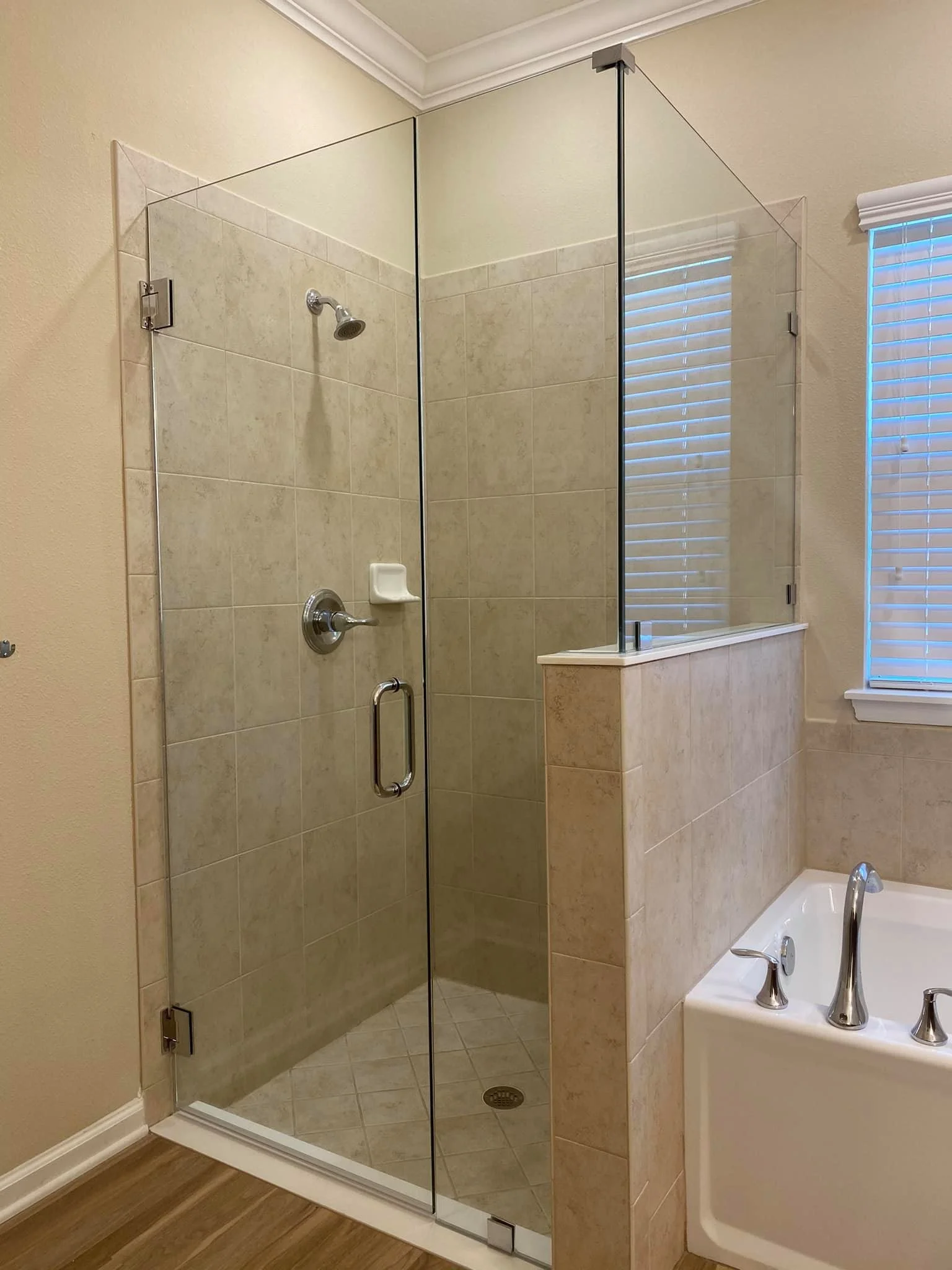 A bathroom featuring a glass-enclosed shower with beige tiled walls and a silver showerhead, positioned next to a large bathtub with a silver faucet, and a window with white blinds.