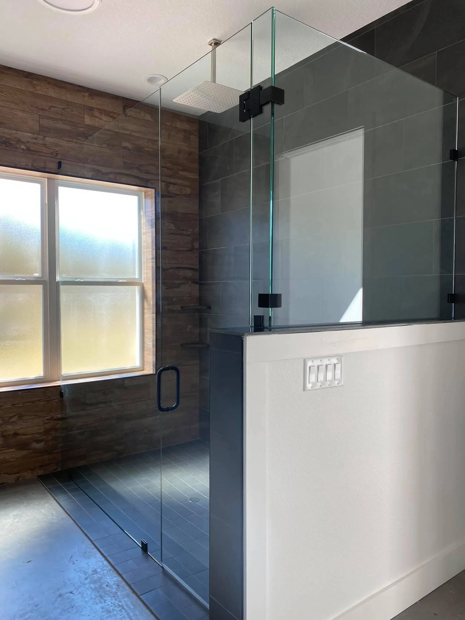 Modern bathroom with walk-in glass shower, wood-paneled wall, frosted window, and white partial wall.