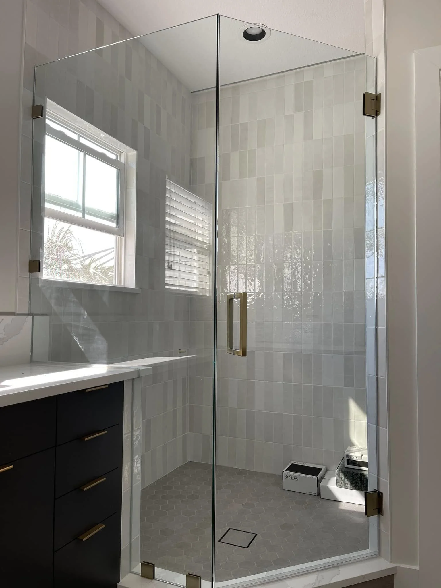 A modern glass shower enclosure with a gold handle, tiled light gray walls, and a small window with blinds, featuring a black overhead showerhead and a corner drain on the mosaic tile floor.