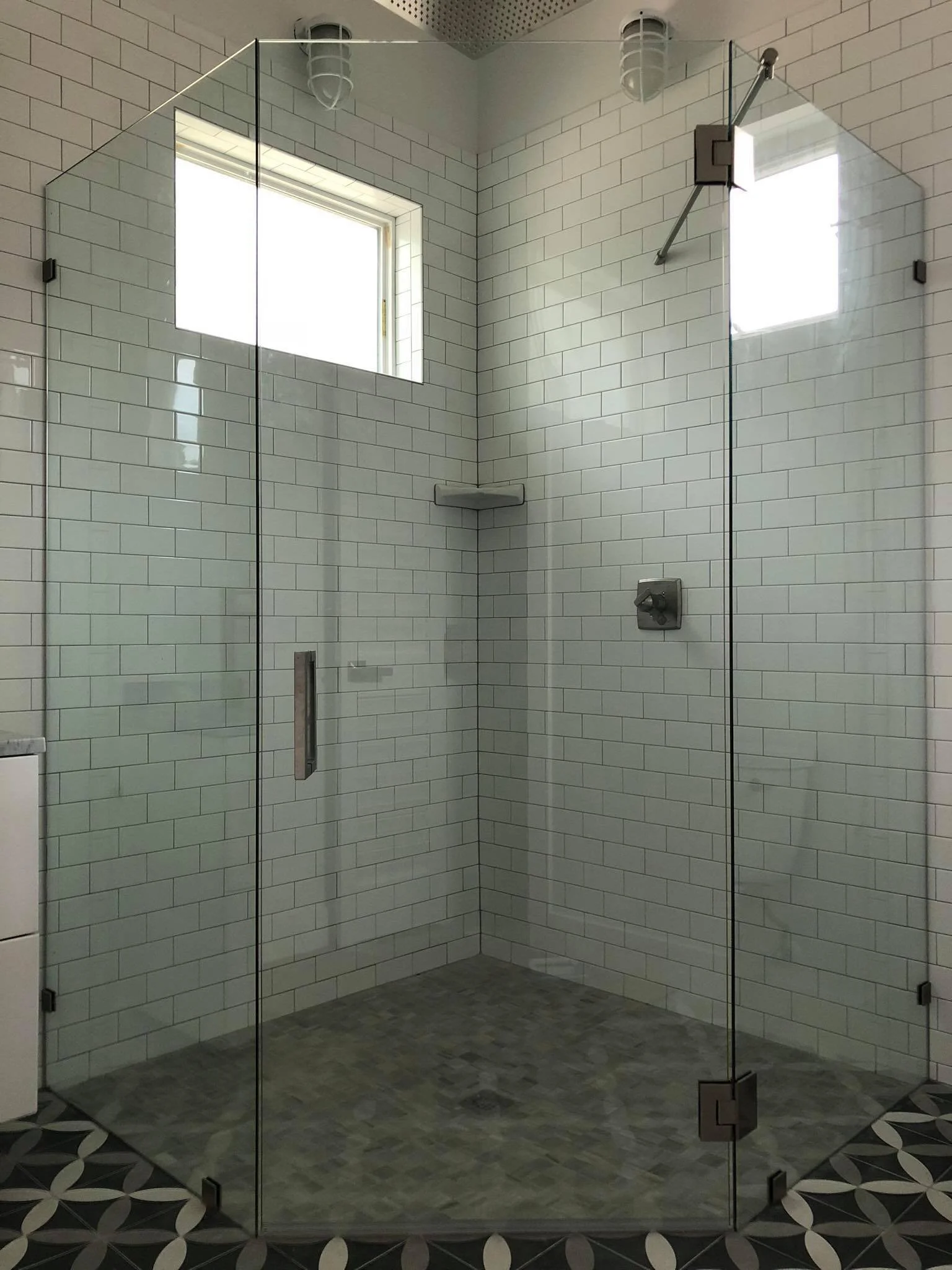 A modern walk-in glass shower with white subway tile walls, a small corner shelf, and a window with natural light, featuring a patterned black and white tile floor.