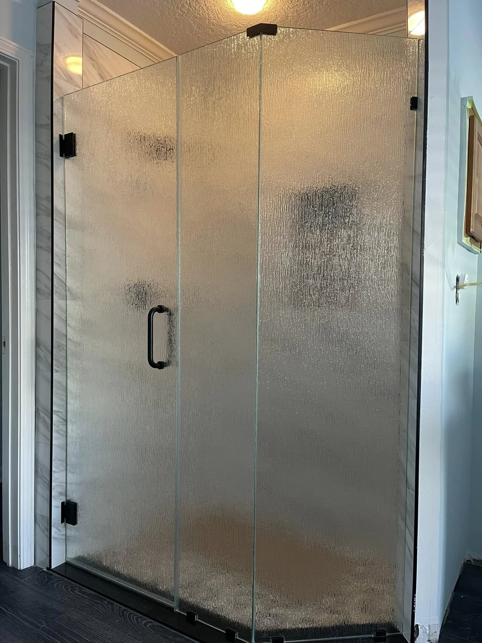 Glass shower door with textured frosted glass and a black handle, framed by white walls and adjacent to a mirror and bathroom fixtures.