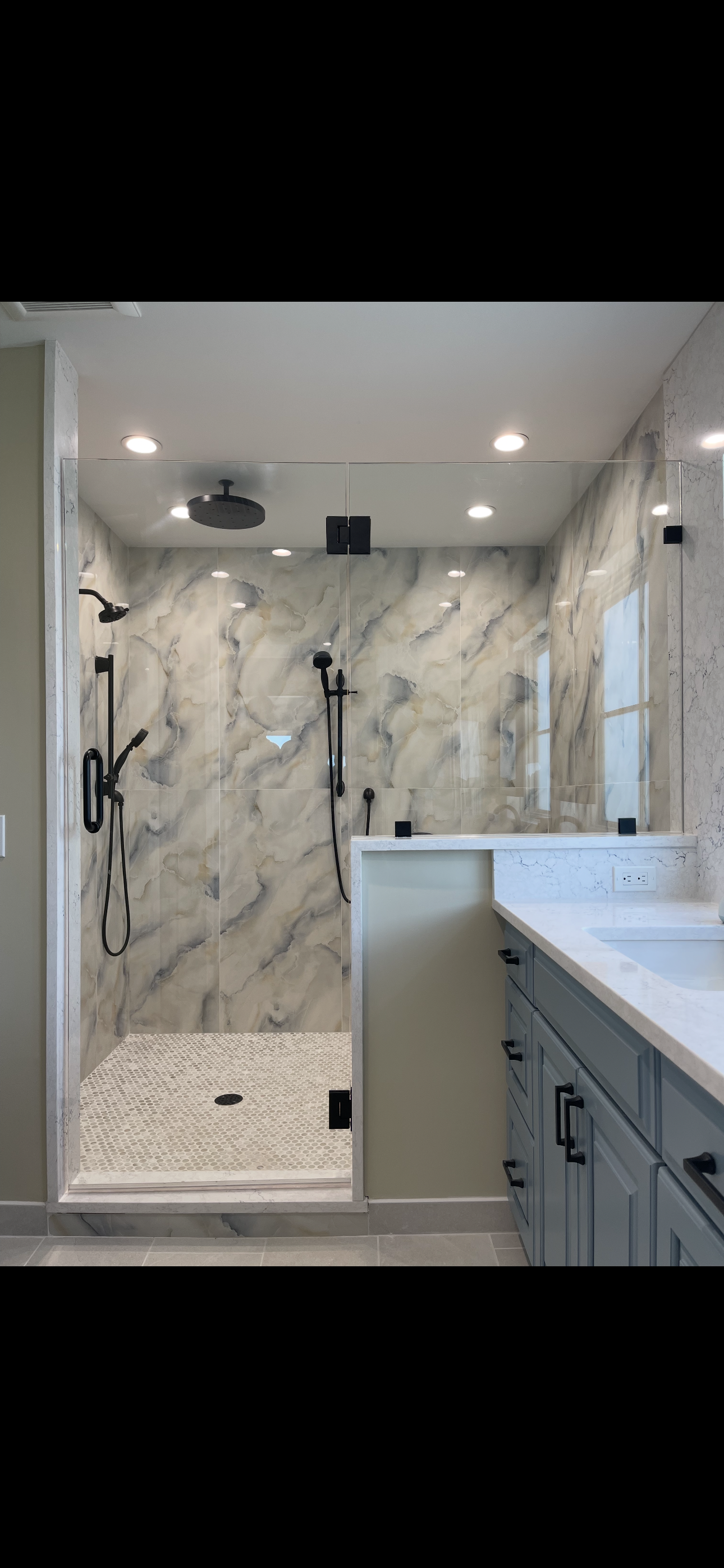 Modern walk-in shower with marble walls, black fixtures, and a glass door, adjacent to a bathroom vanity with a marble countertop and grey cabinets.