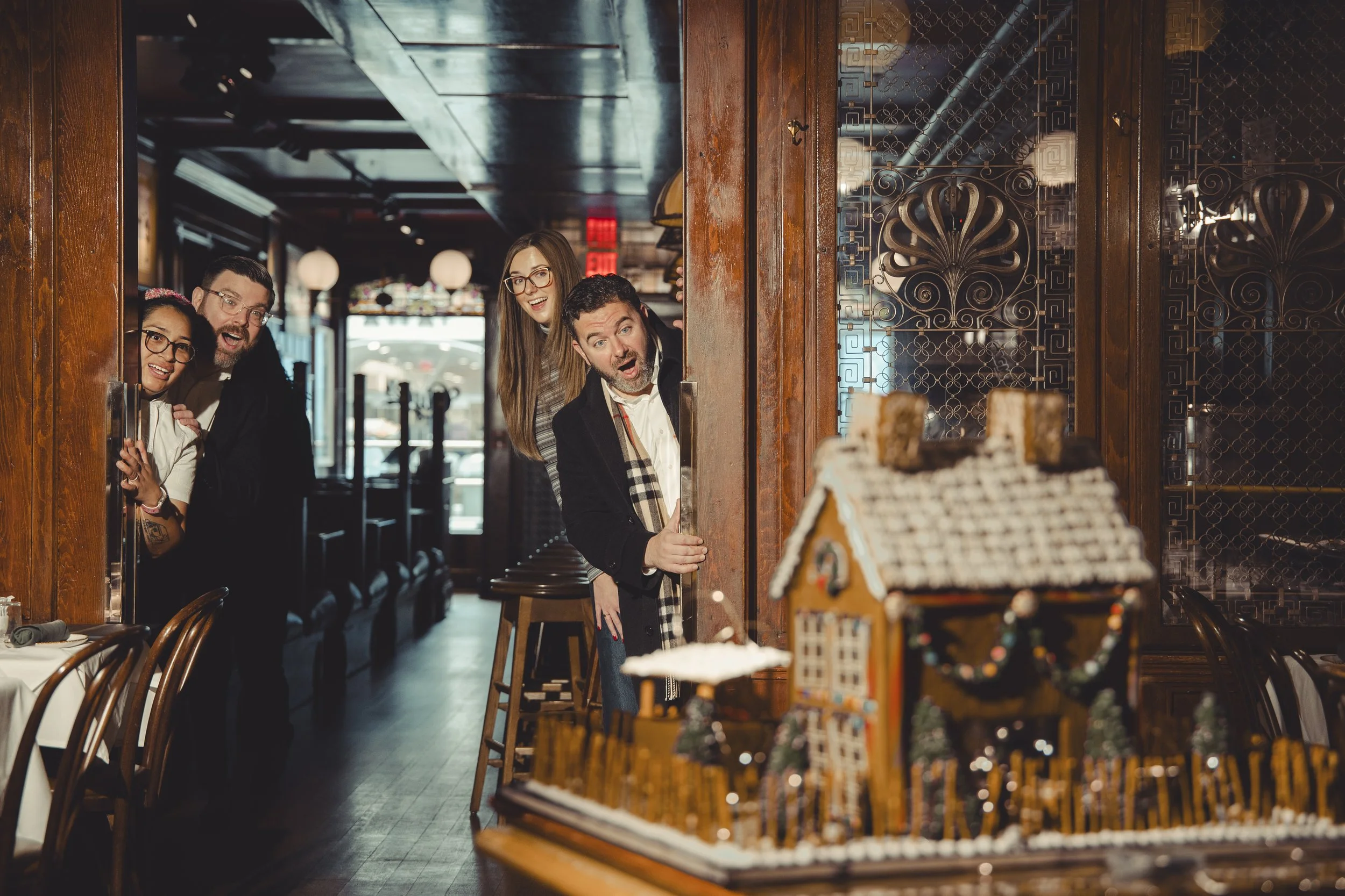 Four people peeking through a doorway and showing surprise at a gingerbread house on a table inside a restaurant.