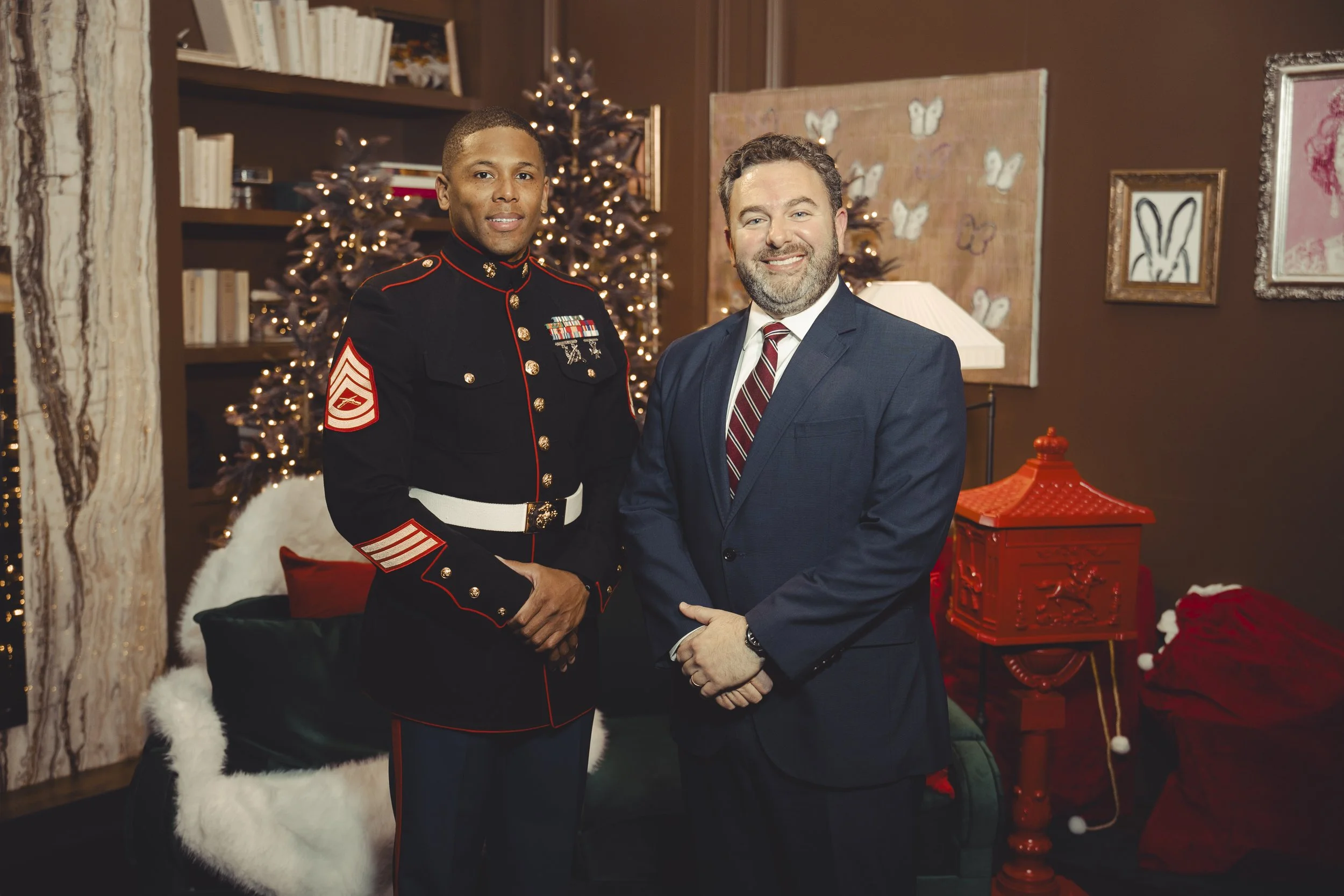 A man in a dark military uniform with medals and stripes standing next to a man in a navy suit with a striped tie. They are smiling and standing in a warmly decorated room with Christmas trees, framed artwork, a red decorative box, and holiday decorations.