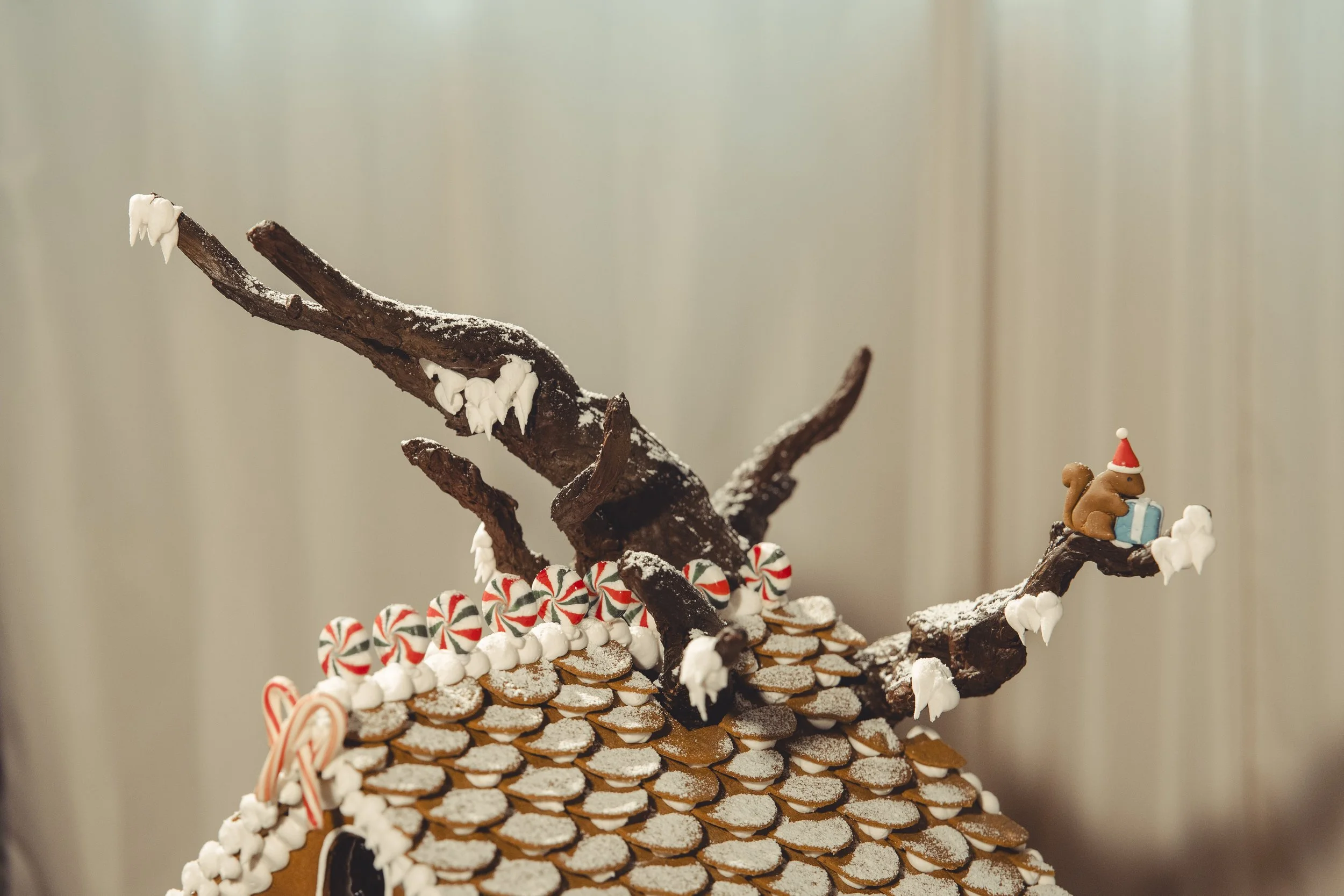 Gingerbread house decorated with peppermint candies, snow-like icing, and chocolate sticks arranged to resemble tree branches with a small squirrel figure wearing a Santa hat on a branch holding a gift.