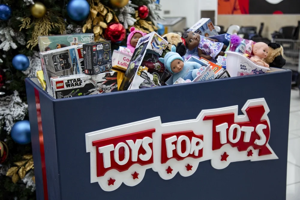 A donation bin filled with toys including dolls, LEGO sets, and toy boxes, with Christmas decorations and a decorated tree in the background.