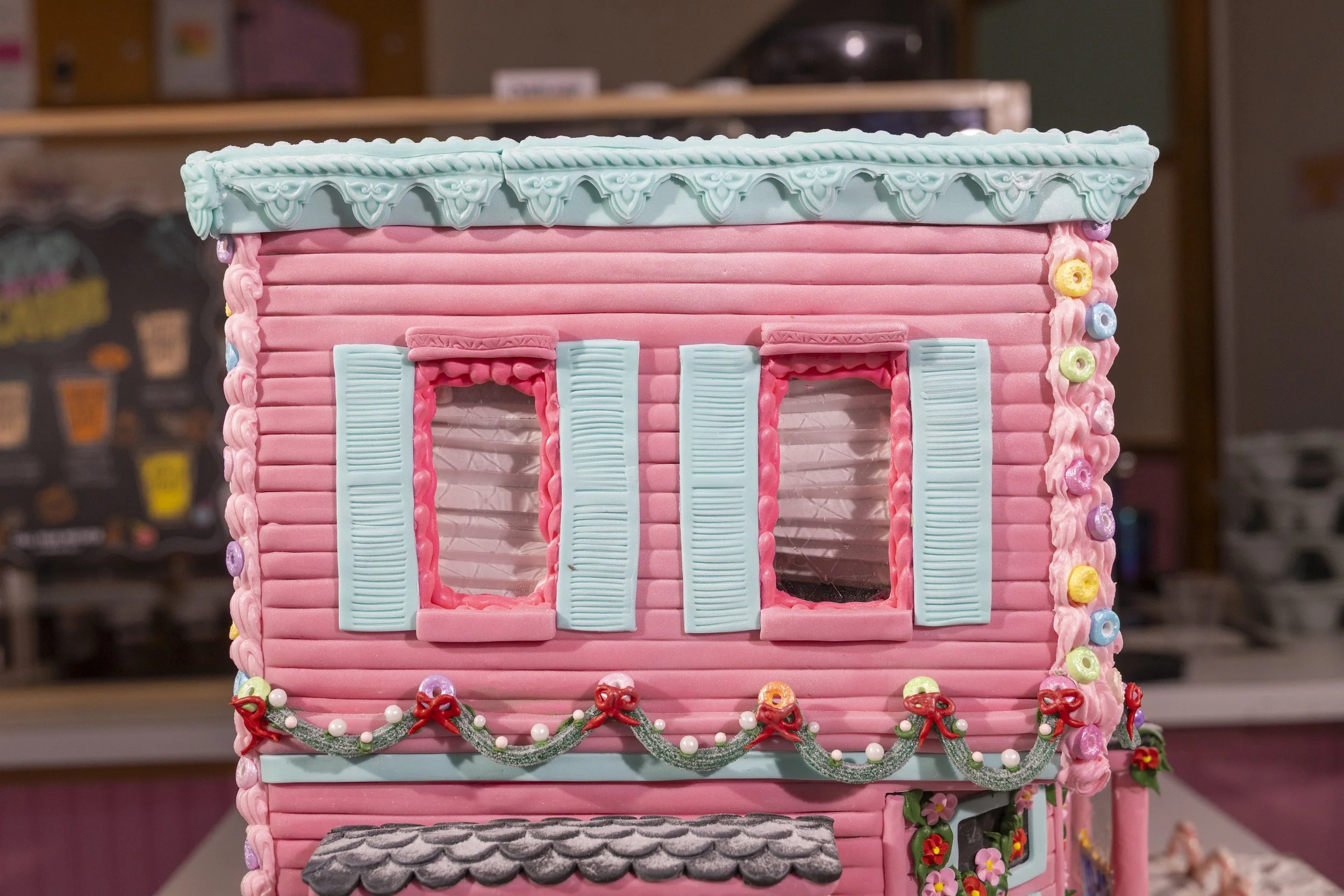 A pink gingerbread house with blue shutters, decorated with colorful candies and festive garlands.
