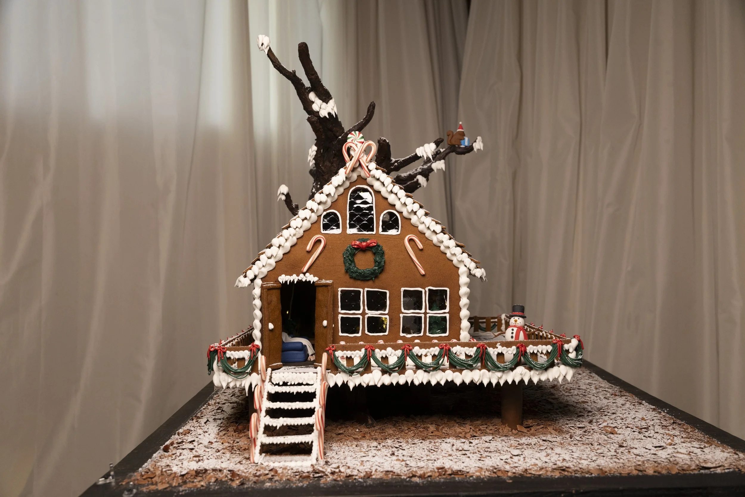Gingerbread house decorated for Christmas with white icing, a small snowman, and a tree with snow on the roof and 