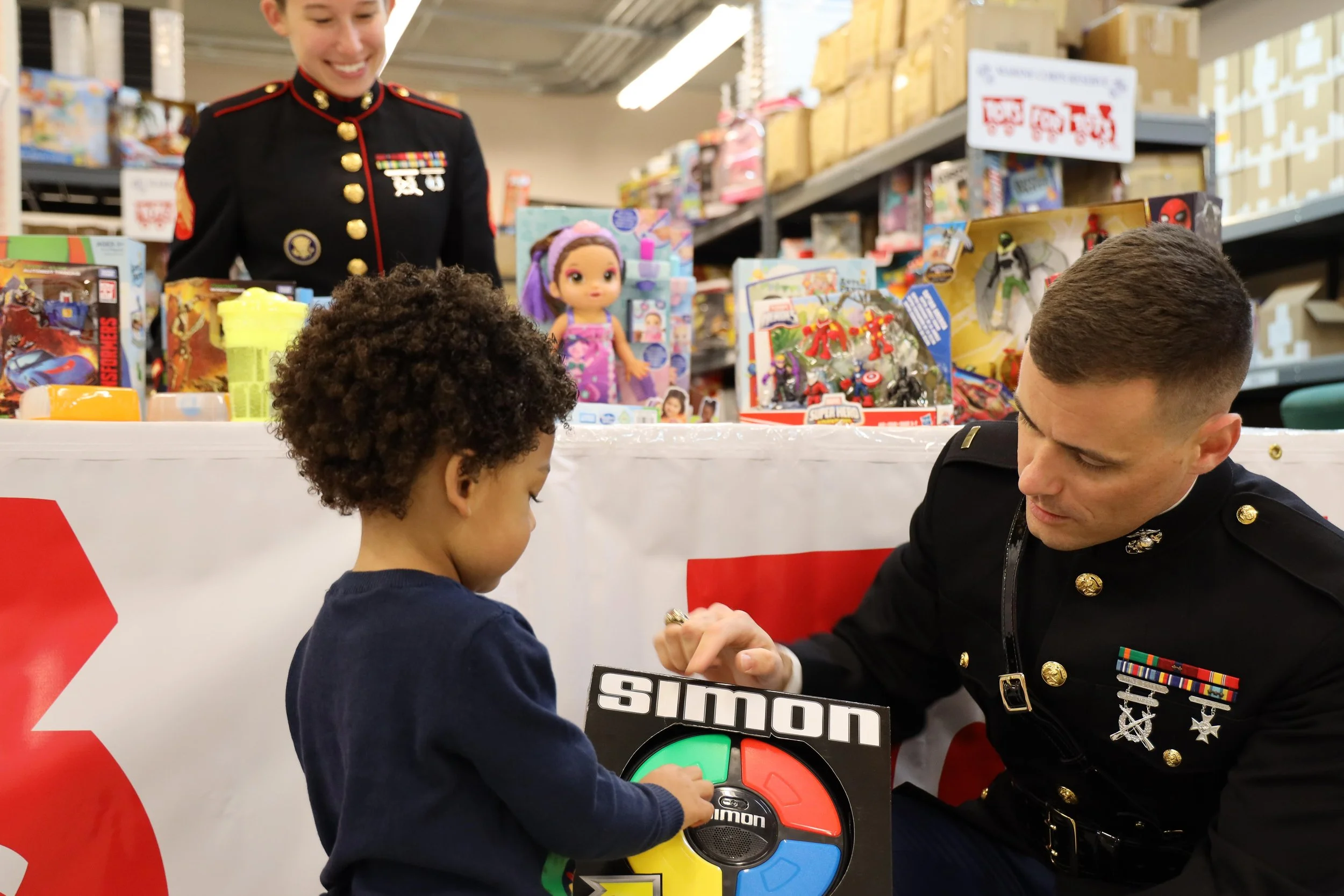 A young boy with curly hair receiving an autograph from a man in a military uniform at a toy giveaway event, with a woman in uniform smiling in the background. Toys and boxed figures are displayed on a table behind them.