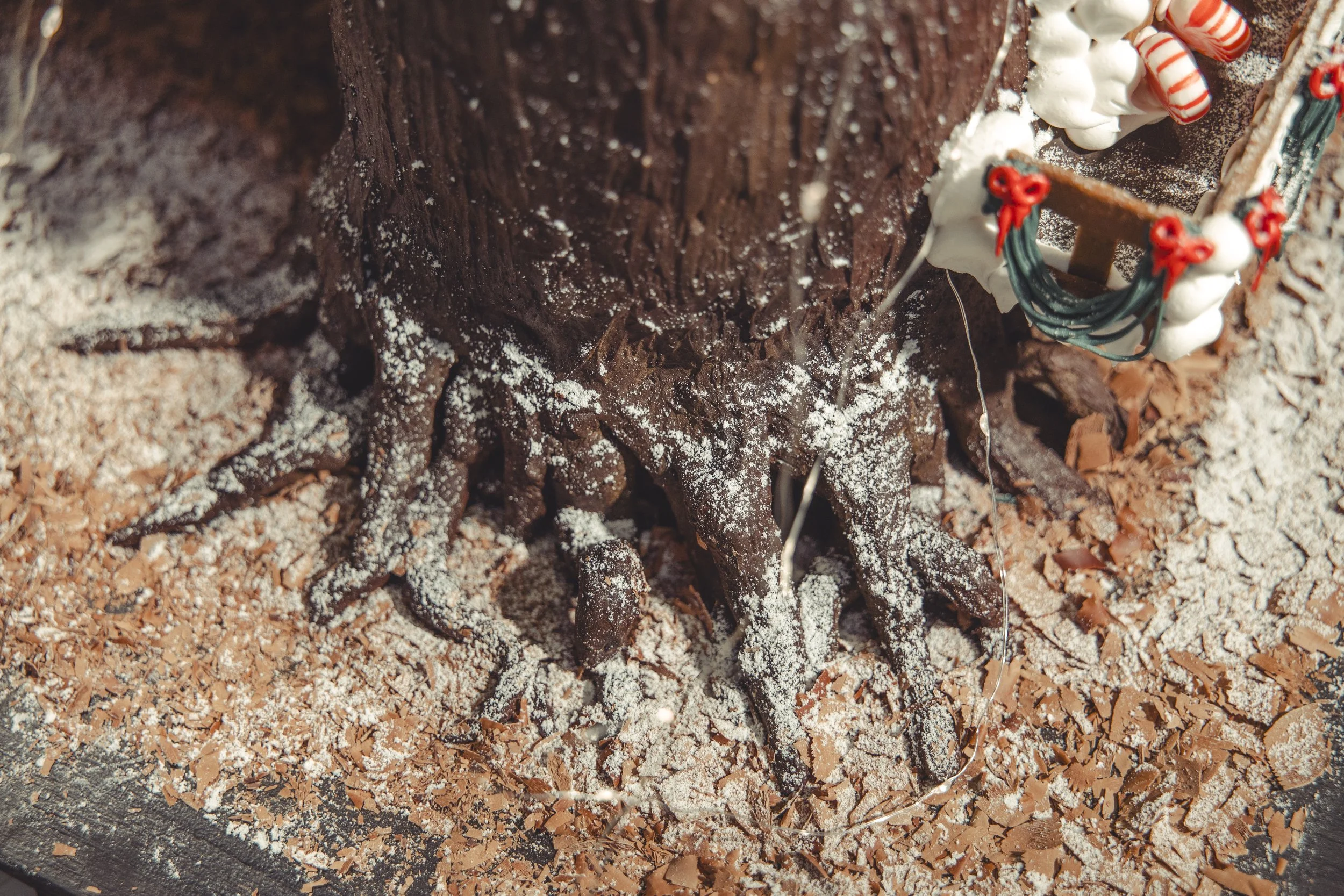 Close-up of a decorated Christmas-themed cake resembling a tree trunk with branches, dusted with powdered sugar, on a surface scattered with chocolate shavings and sprinkles.