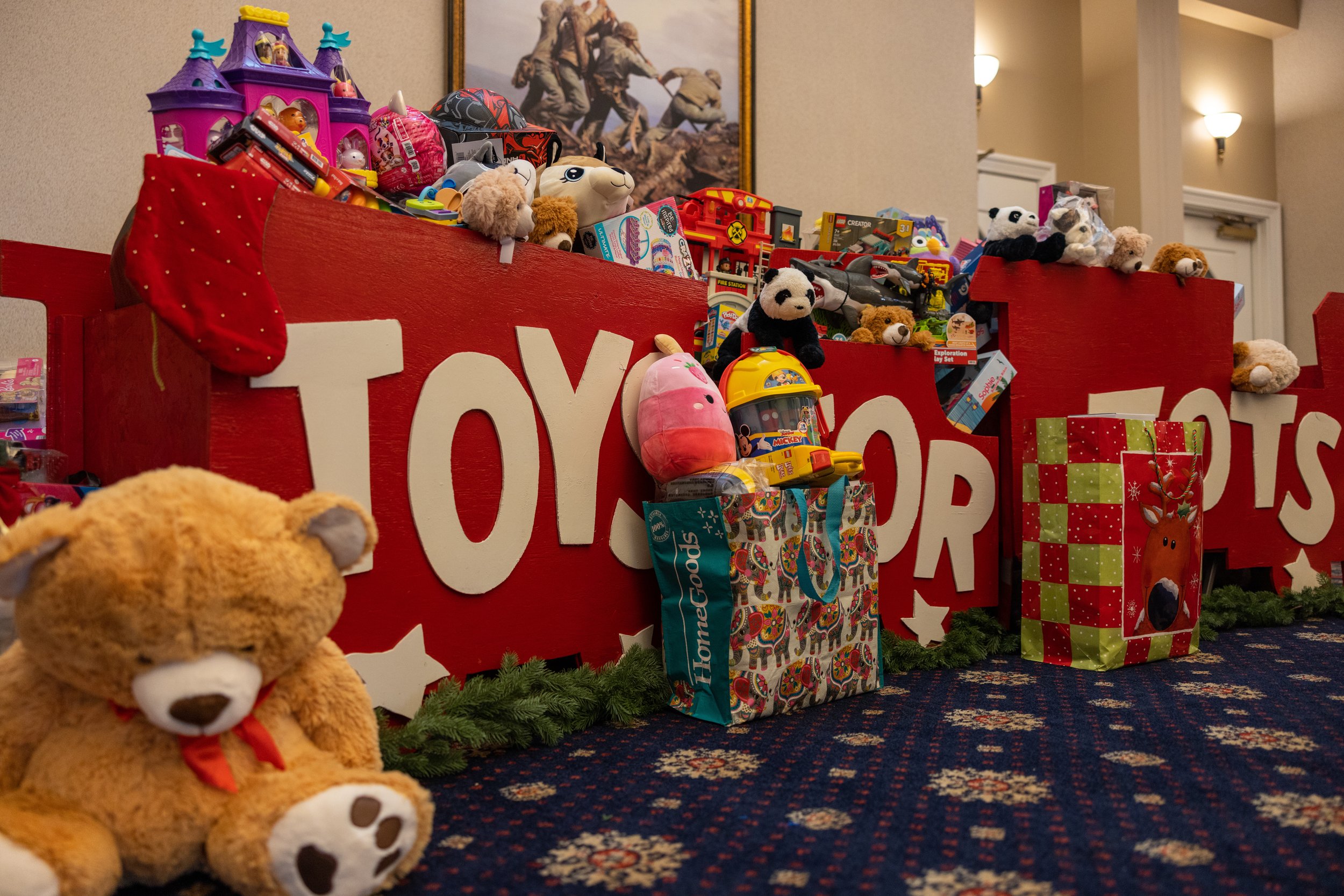 A Christmas display of toys and gifts with large red signs spelling 'TOYS' and 'GIFTS,' including plush teddy bears, toy cars, and wrapped presents, arranged on a carpeted floor in front of a wall with reindeer and winter decor.