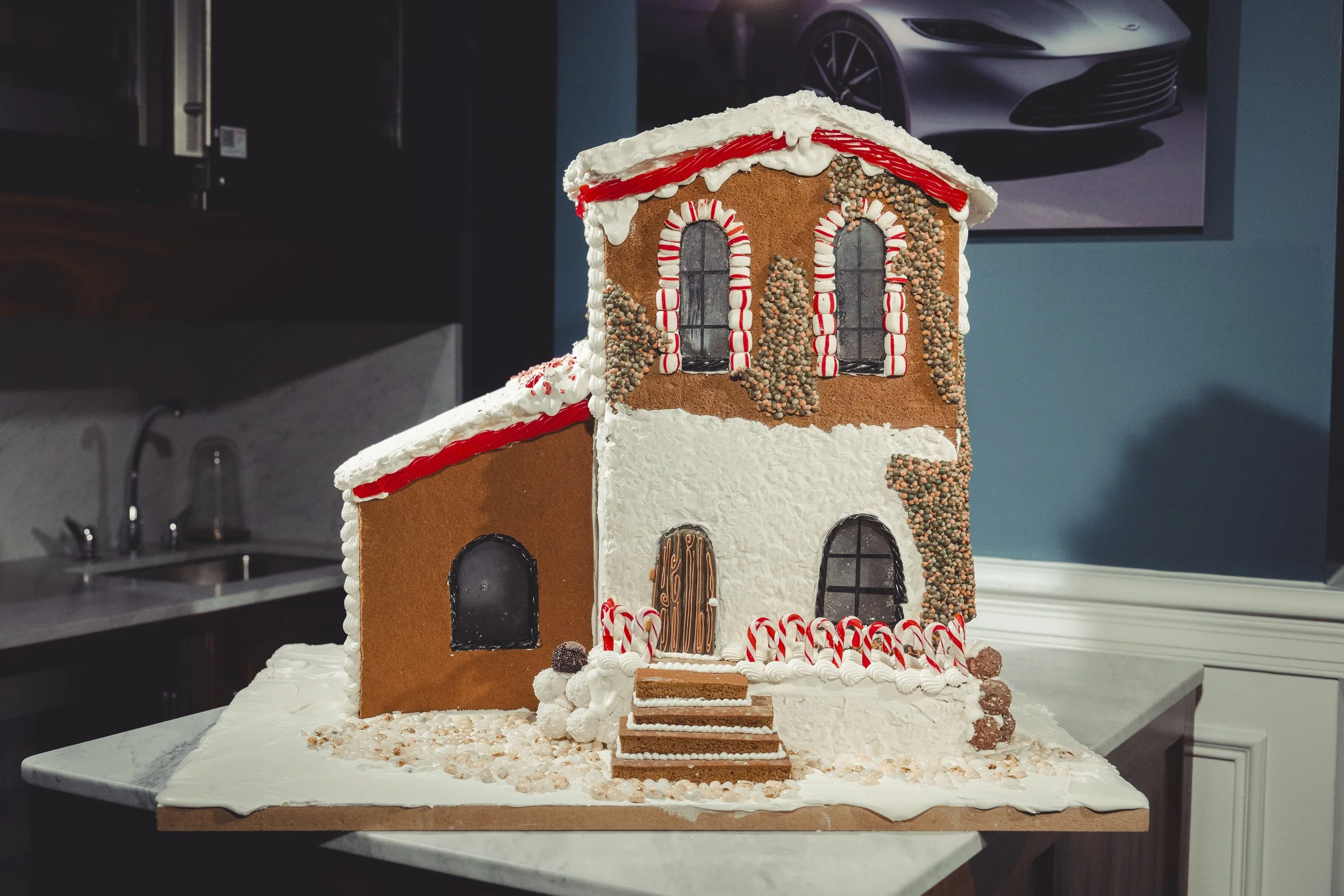 Gingerbread house decorated with white, red, and brown icing, candy canes, and gumdrops, placed on a countertop in a kitchen.
