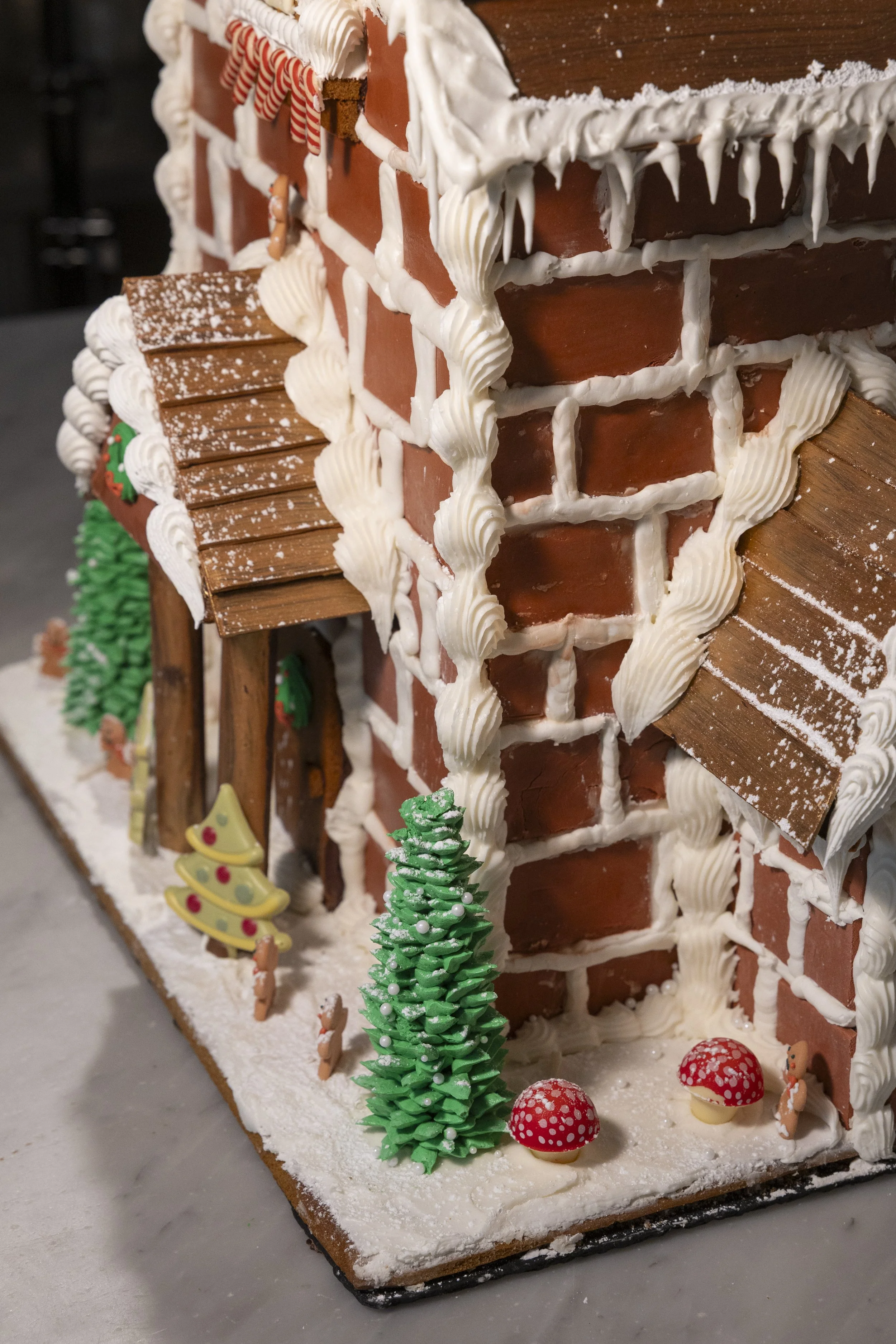 Gingerbread house decorated with icing, candy, and frosting, featuring a brick chimney, trees, and mushrooms, on a snowy base.