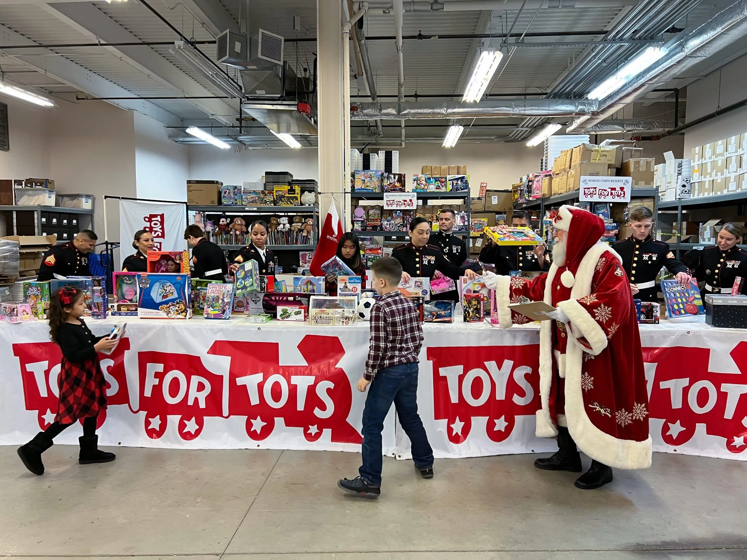 Children and holiday characters, including Santa Claus, at a Toys for Tots toy drive table with volunteers in military uniforms behind it, inside a warehouse filled with shelves of toys and boxes.
