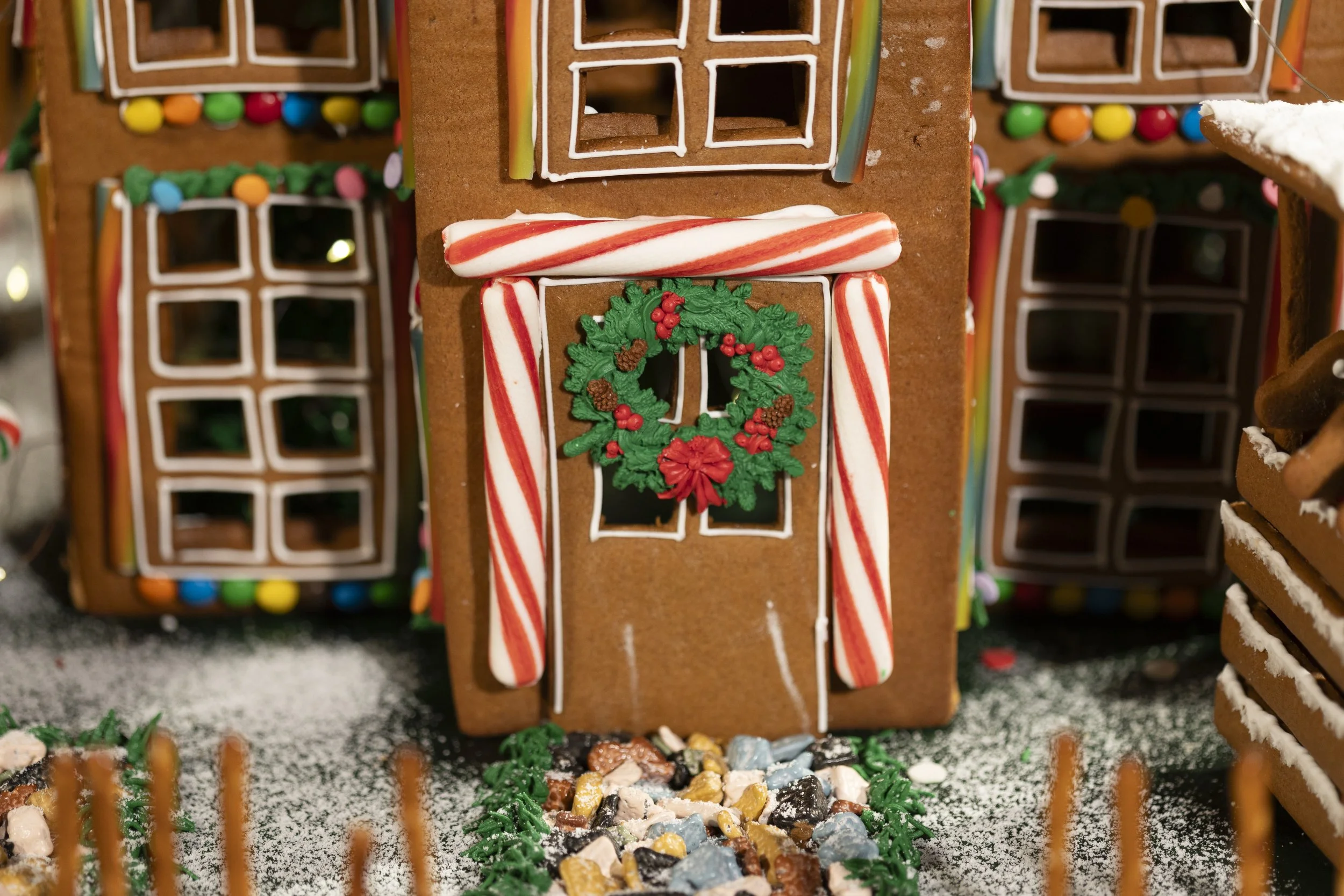 Gingerbread house decorated with peppermint candy canes, a wreath, and colorful candies on the roof, with a path of crushed candies leading to the door, surrounded by a small fence and dusted with powdered sugar.