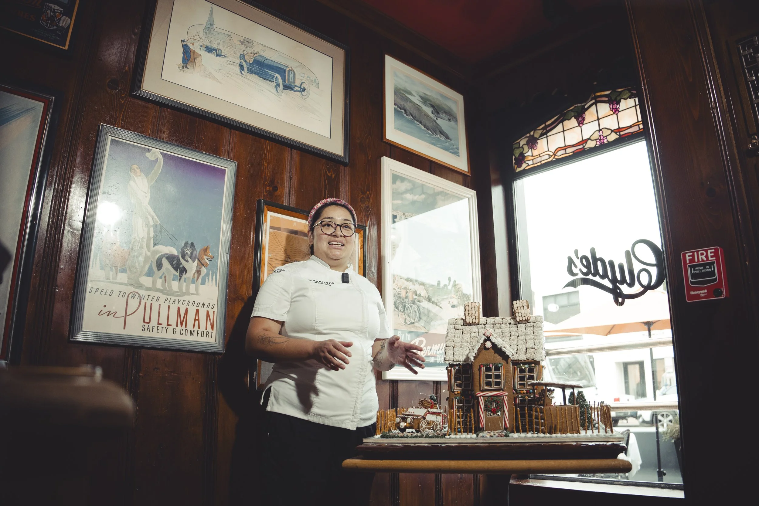 Woman in chef's uniform standing inside a restaurant near a gingerbread house display, with framed pictures on dark wood walls and a large window with stained glass at the top.