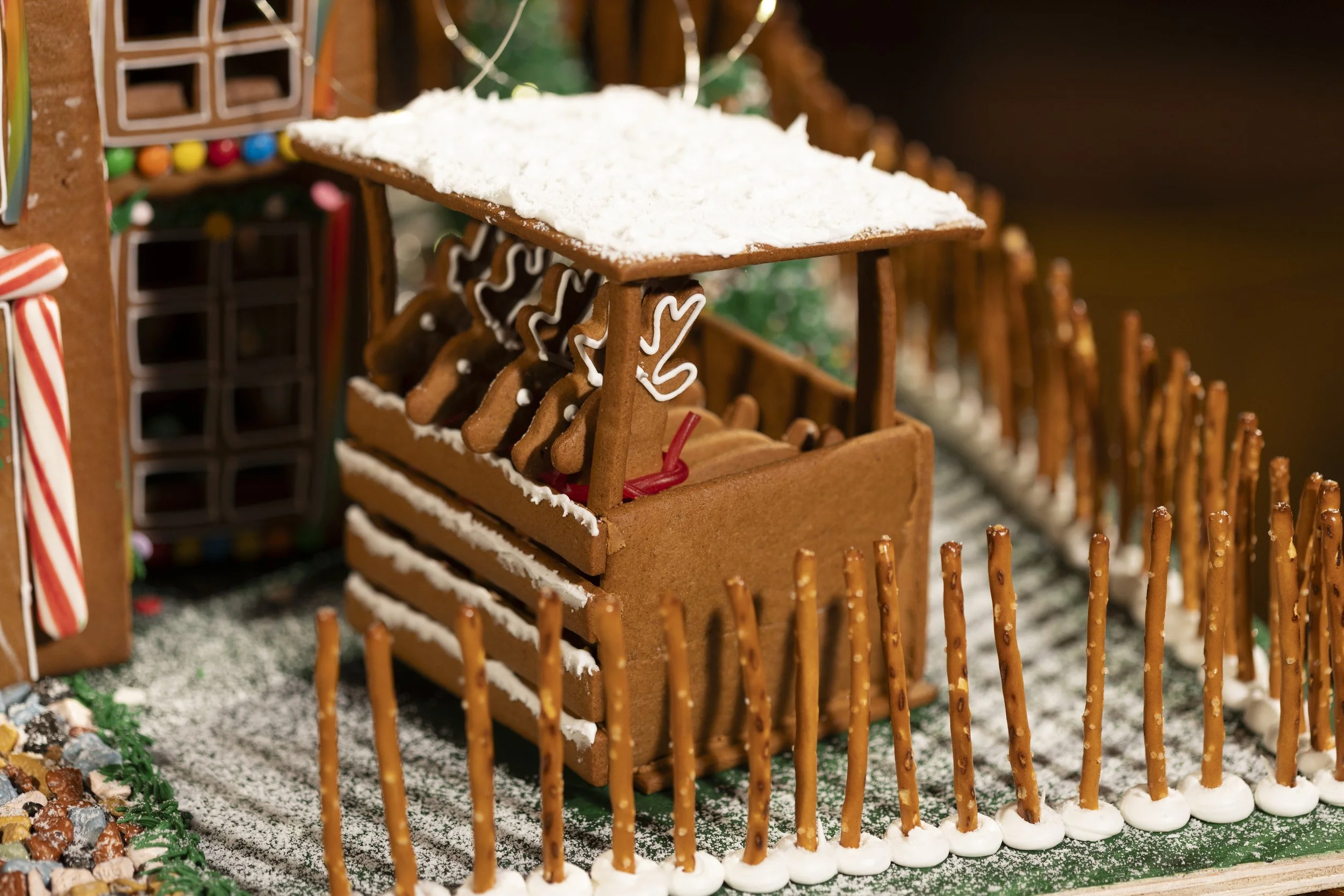 Close-up of a gingerbread house with a small gingerbread wagon filled with gingerbread cookies, surrounded by pretzels and a green base decorated with white icing and powdered sugar.