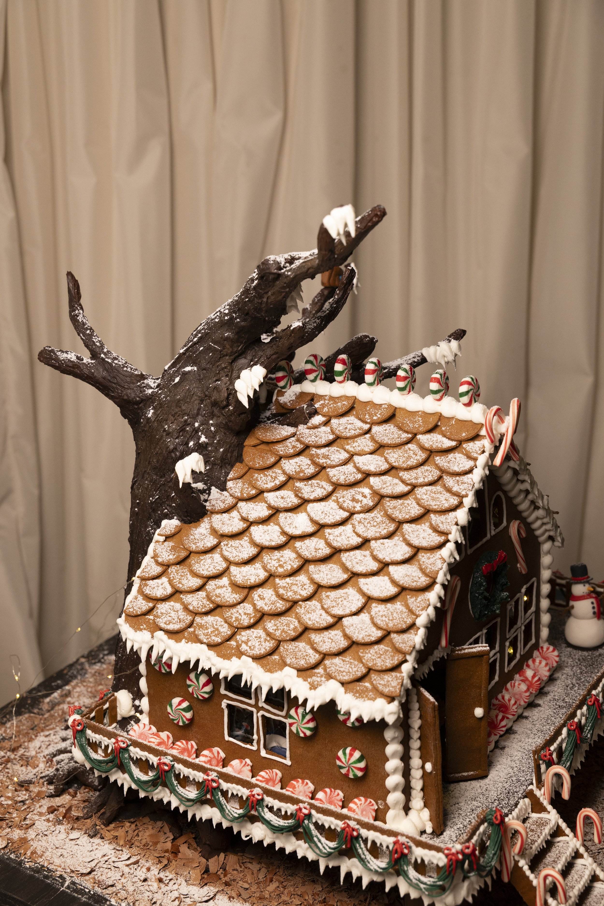 Decorated gingerbread house with a Christmas theme, featuring a brown roof with white powdered sugar, candy cane decorations, a snowman figurine, and a large imitation tree made of dark brown material with white accents.