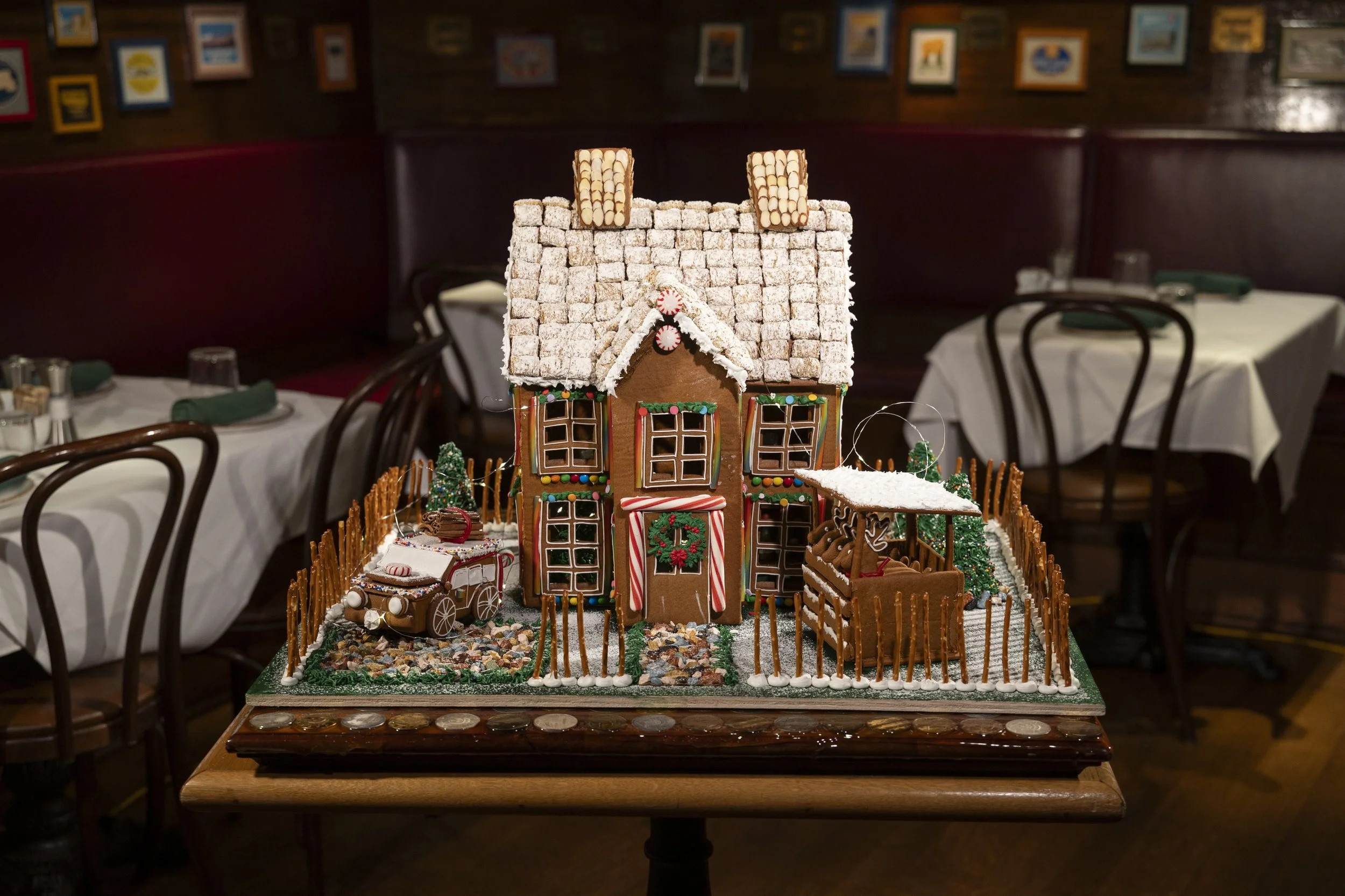 Decorated gingerbread house with a roof covered in powdered sugar, a small car with a wreath on its hood, and a fenced yard with miniature Christmas trees inside an elegant restaurant.
