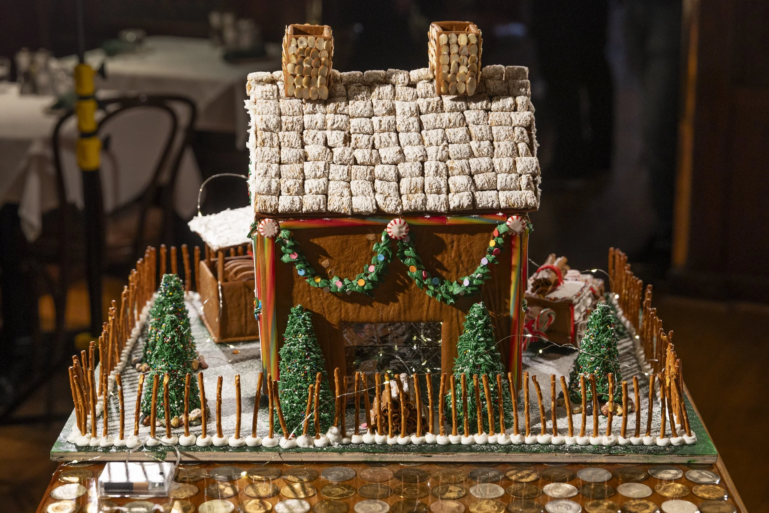A gingerbread house decorated with icing, candies, and miniature Christmas trees, surrounded by a fence made of pretzels, with a snowy yardscape and chocolate coins on the table.