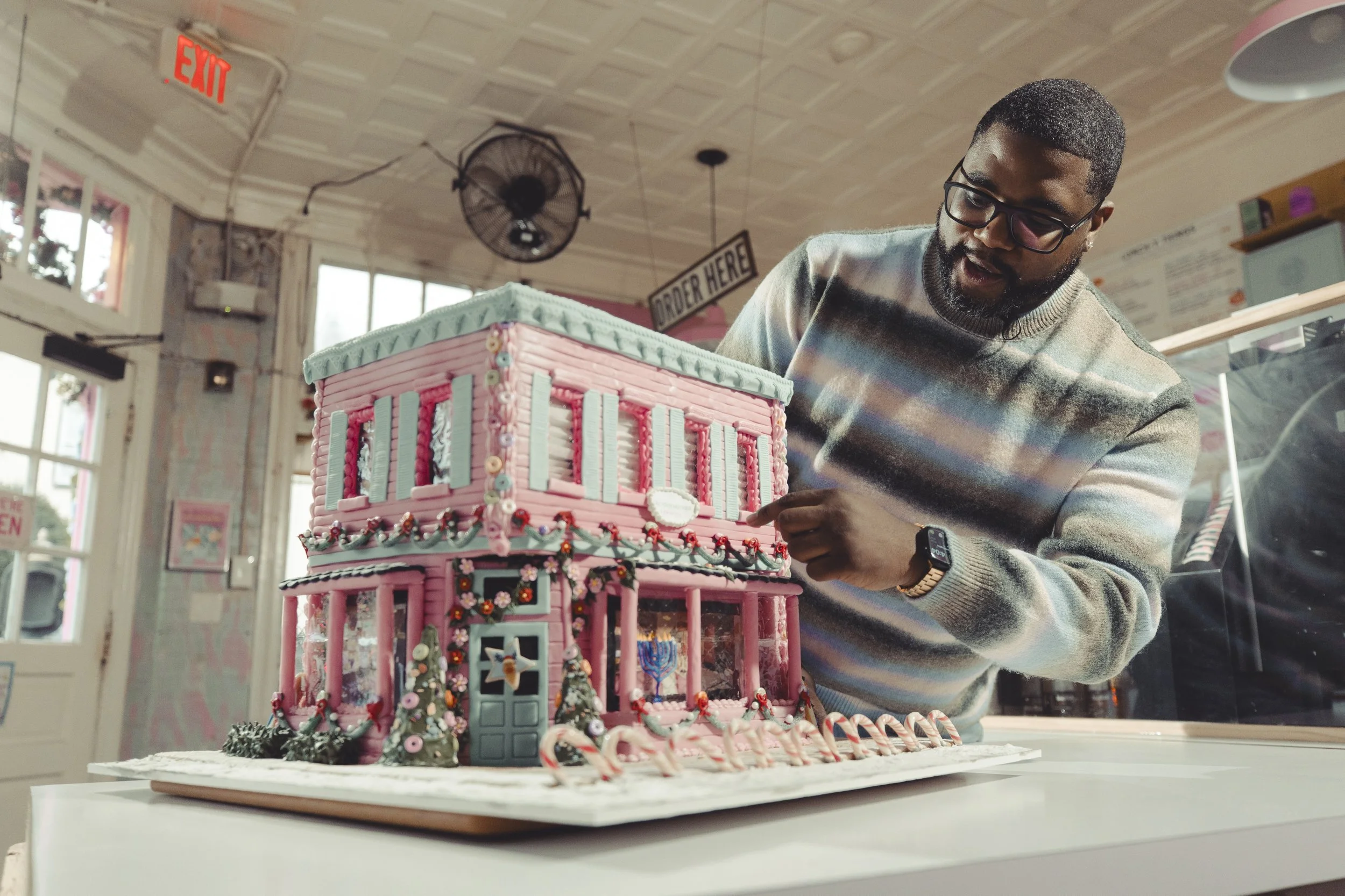 A man in glasses and a striped sweater points to a pink and blue gingerbread house decorated with candies and Christmas ornaments inside a bakery or cafe.