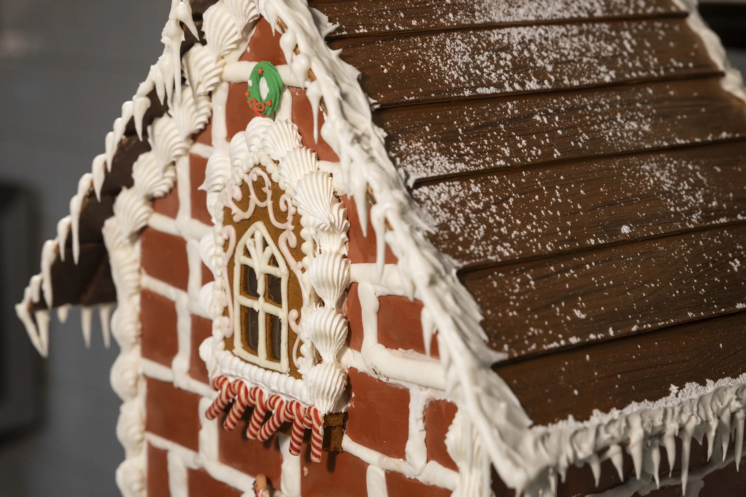 Close-up of a gingerbread house with white icing, decorated with peppermint candies and a small Christmas wreath ornament, set against a brown wooden background dusted with powdered sugar.