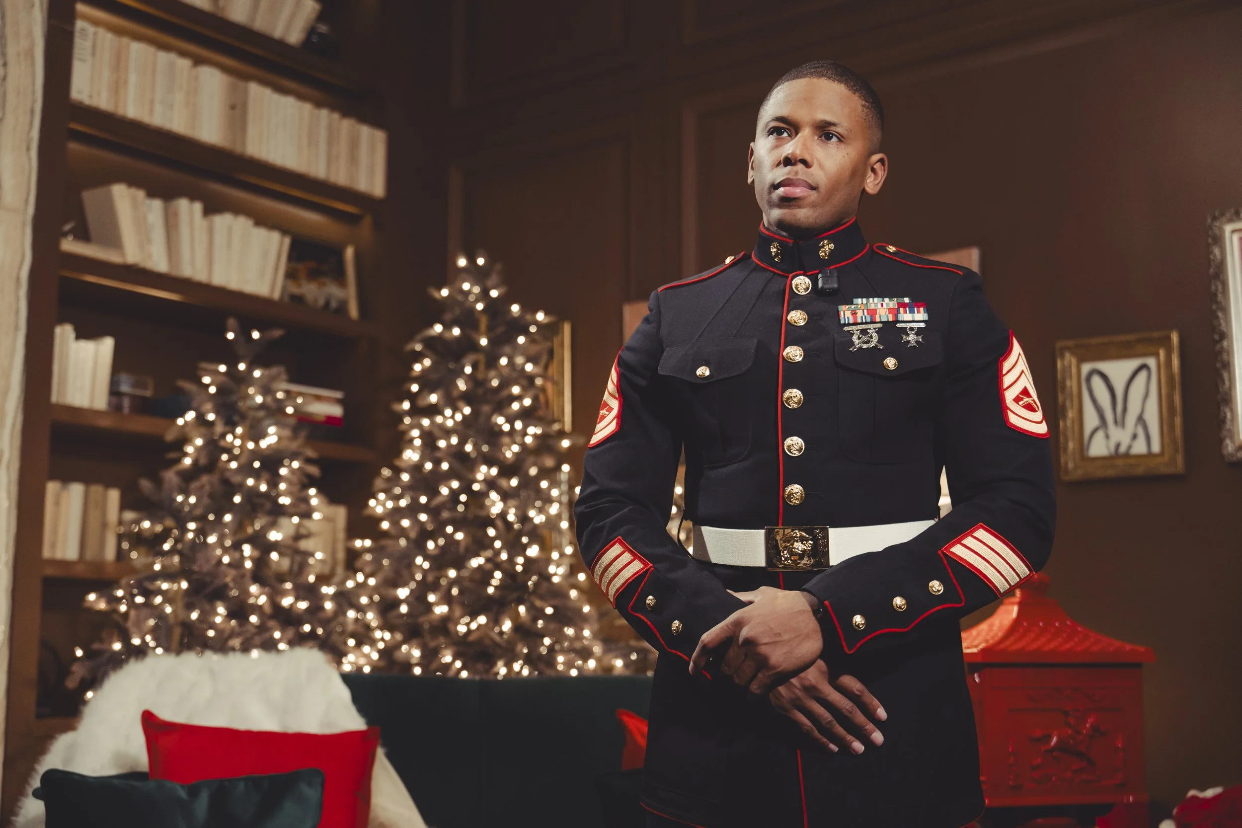 A man in a U.S. Marine dress uniform standing in a room decorated for Christmas, with Christmas trees with lights, a bookshelf, and holiday-themed decorations in the background.