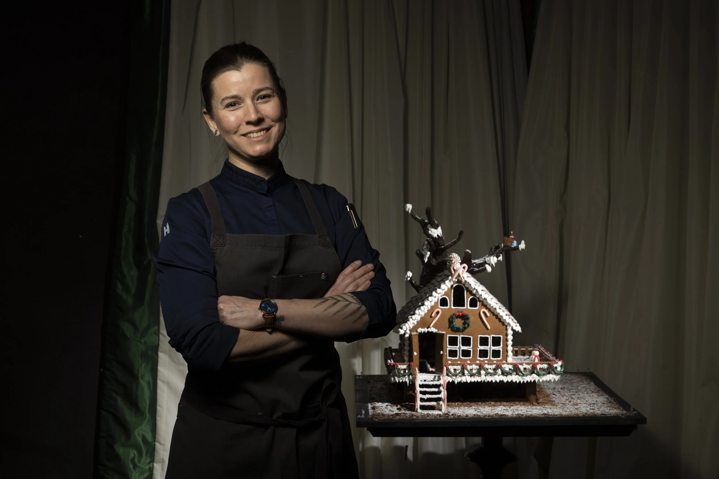 A woman with crossed arms standing next to a decorated gingerbread house, smiling at the camera in a dark setting with curtains in the background.