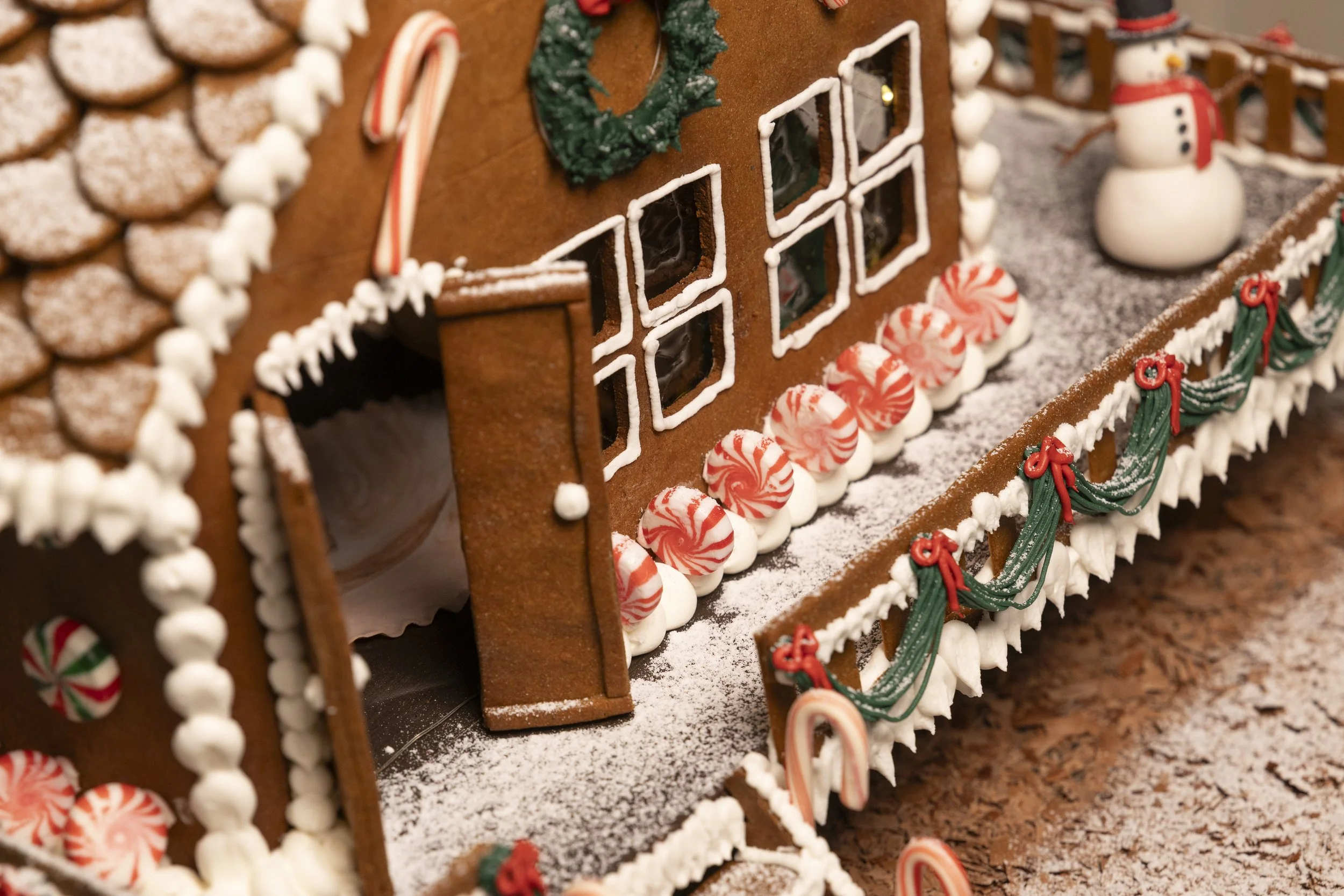 Close-up of a decorated gingerbread house with peppermint candies, green icing garlands, a snowman figure, and candy canes on a snowy surface.